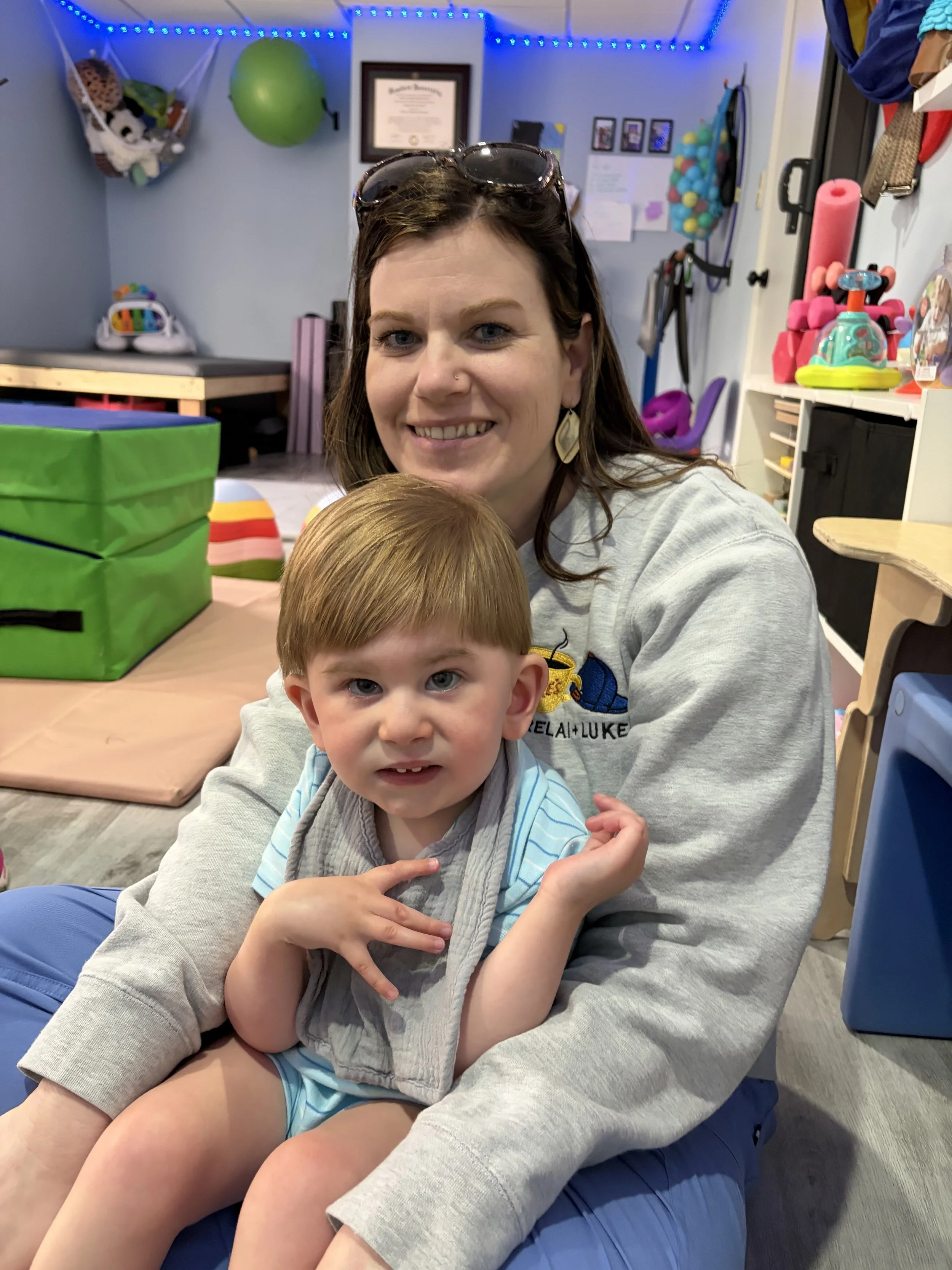 A woman with brown hair, sunglasses on her head, smiling, sitting next to a young boy with red hair and blue eyes. They are indoors with toys and colorful decorations in the background.