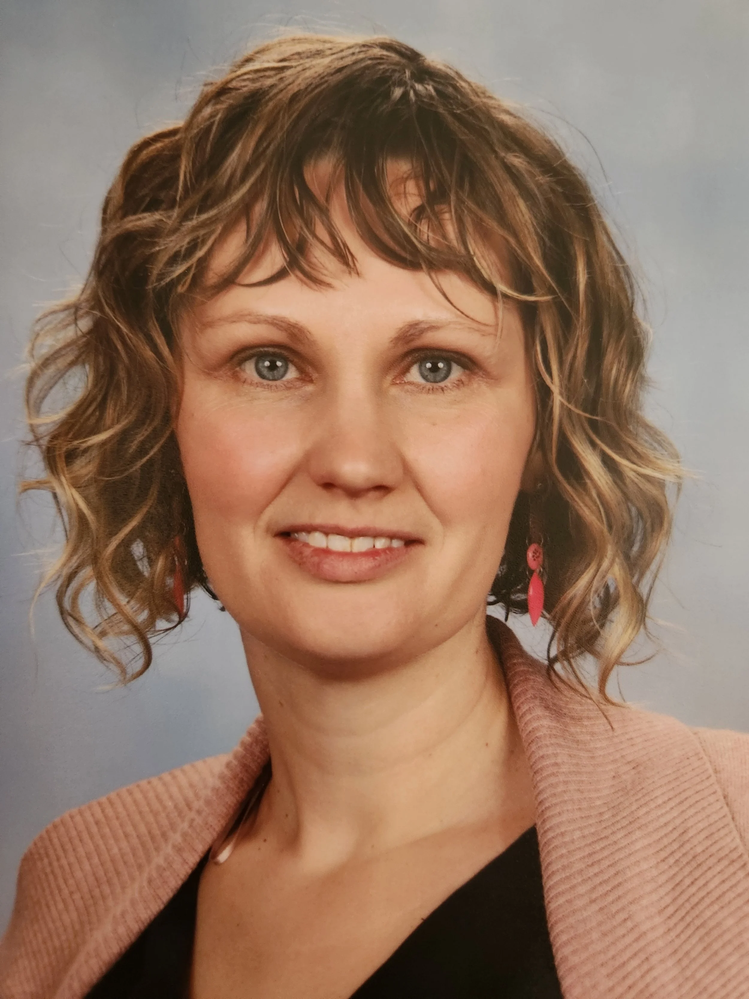 A woman with curly, light brown hair and blue eyes, wearing a pink sweater and earrings, smiling against a gray background.