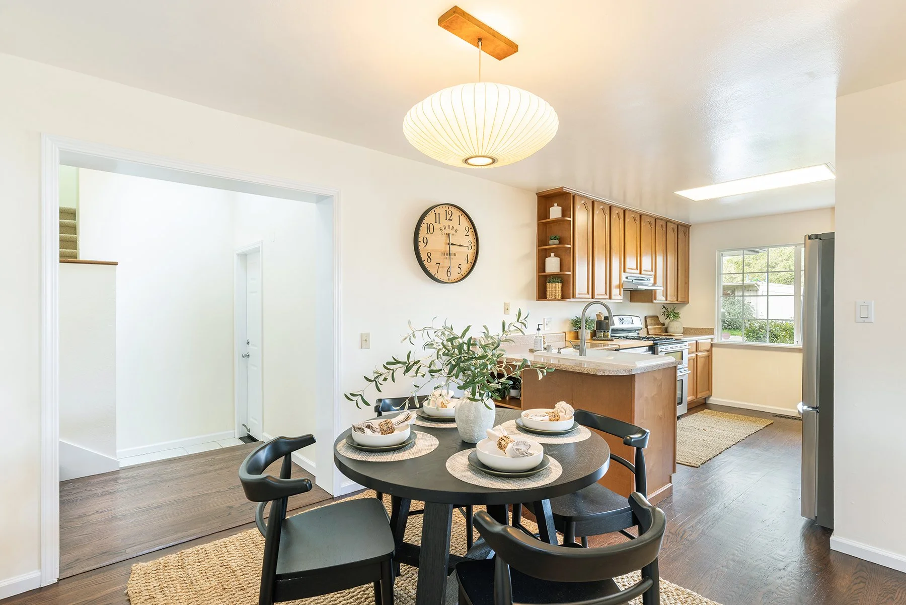 Inside a cozy kitchen with a round dining table set for four, a large wall clock, wooden cabinets, granite countertops, stainless steel appliances, and a window with natural light.