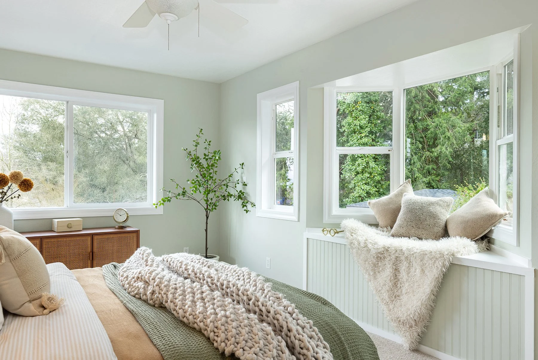 Bright bedroom with large windows overlooking greenery, cozy bed with chunky knit blanket, beige and cream pillows, a small wooden cabinet, a potted plant, and decorative items on the window ledge.