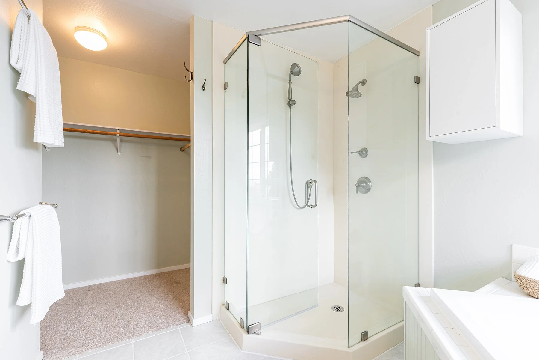 A bathroom with a glass shower stall, a white cabinet on the wall, and a walk-in closet with a wooden rod and shelf.