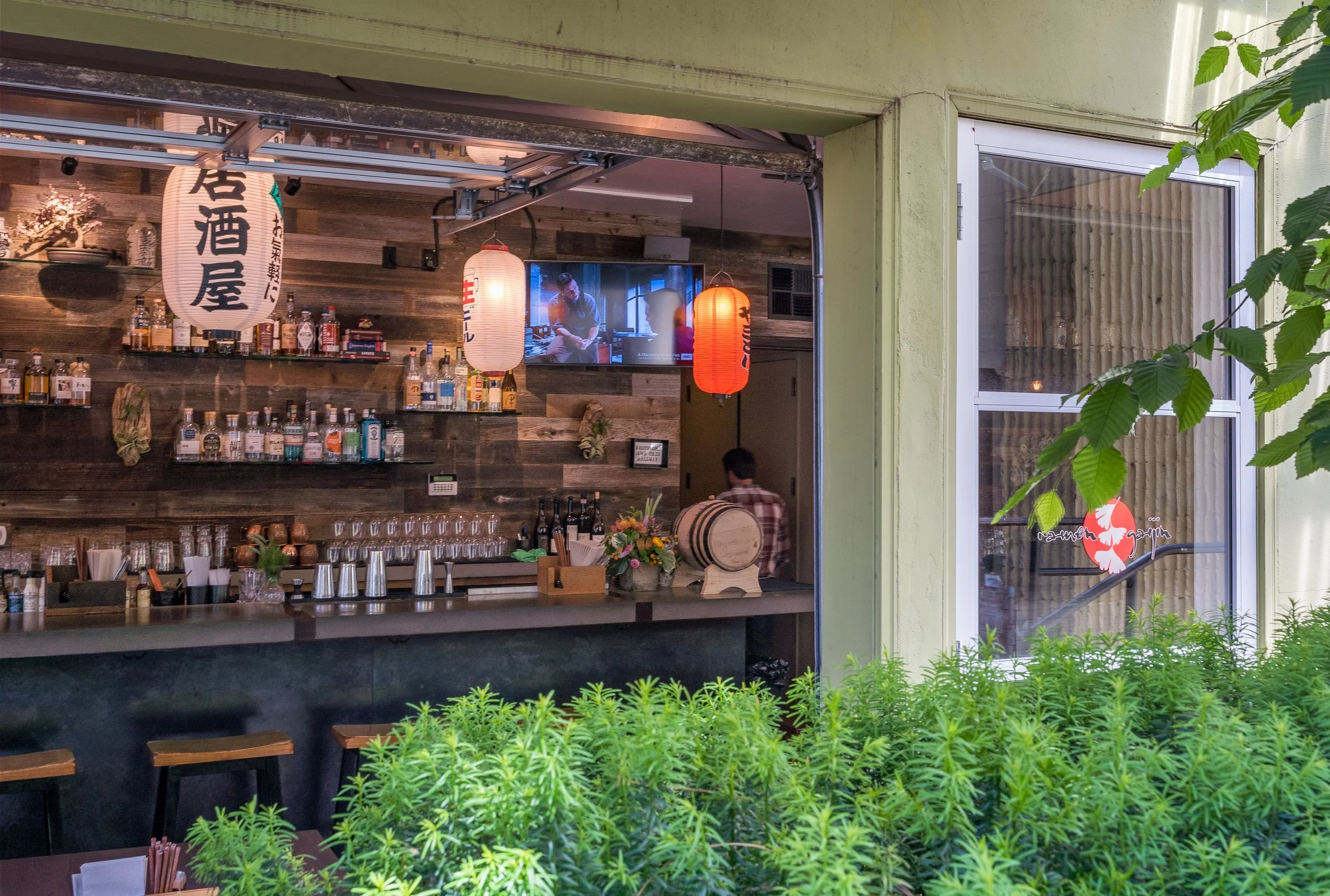 View of a cozy Japanese-style bar with wooden shelves stocked with alcohol bottles, hanging paper lanterns, a TV screen, a person working behind the bar, and outside greenery in the foreground.