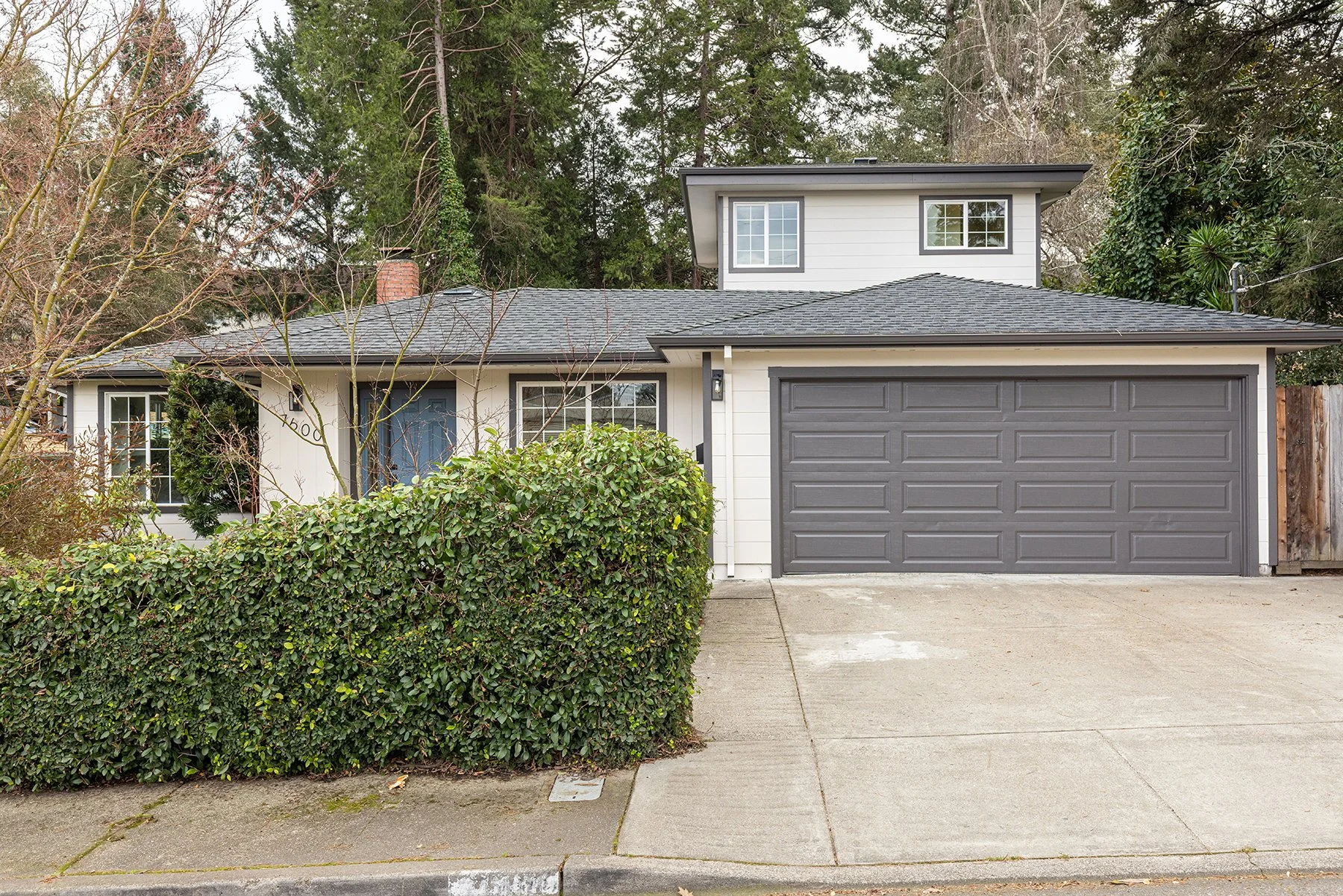 Front view of a two-story house with gray garage door, white siding, blue front door, and surrounding greenery, including bushes and trees.
