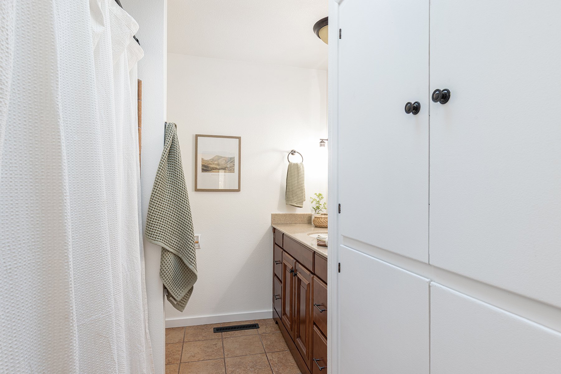 A small bathroom with a wooden vanity, a framed picture on the wall, beige towels on a ring and in a basket, a potted plant, and a white cabinet.