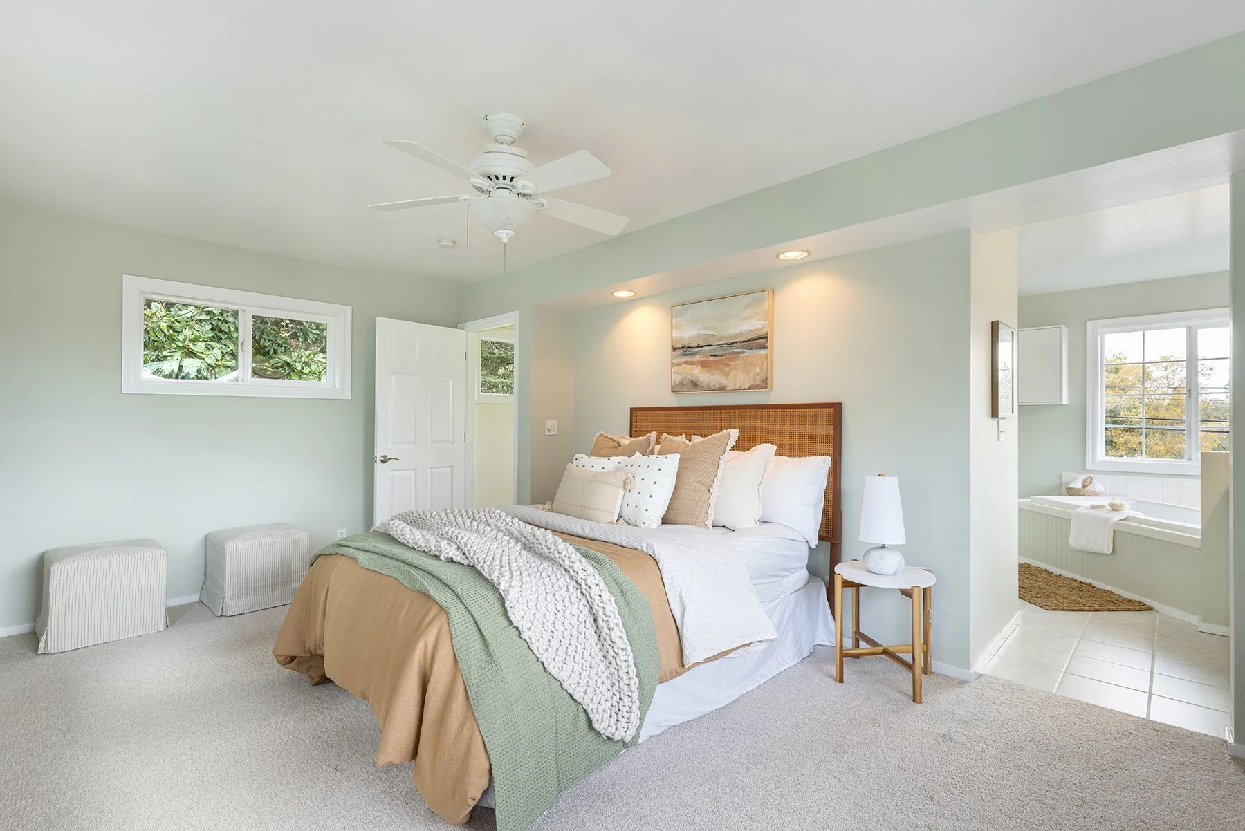 A bedroom with a bed, throw pillows, nightstand with a lamp, small stools, and a window showing trees outside.