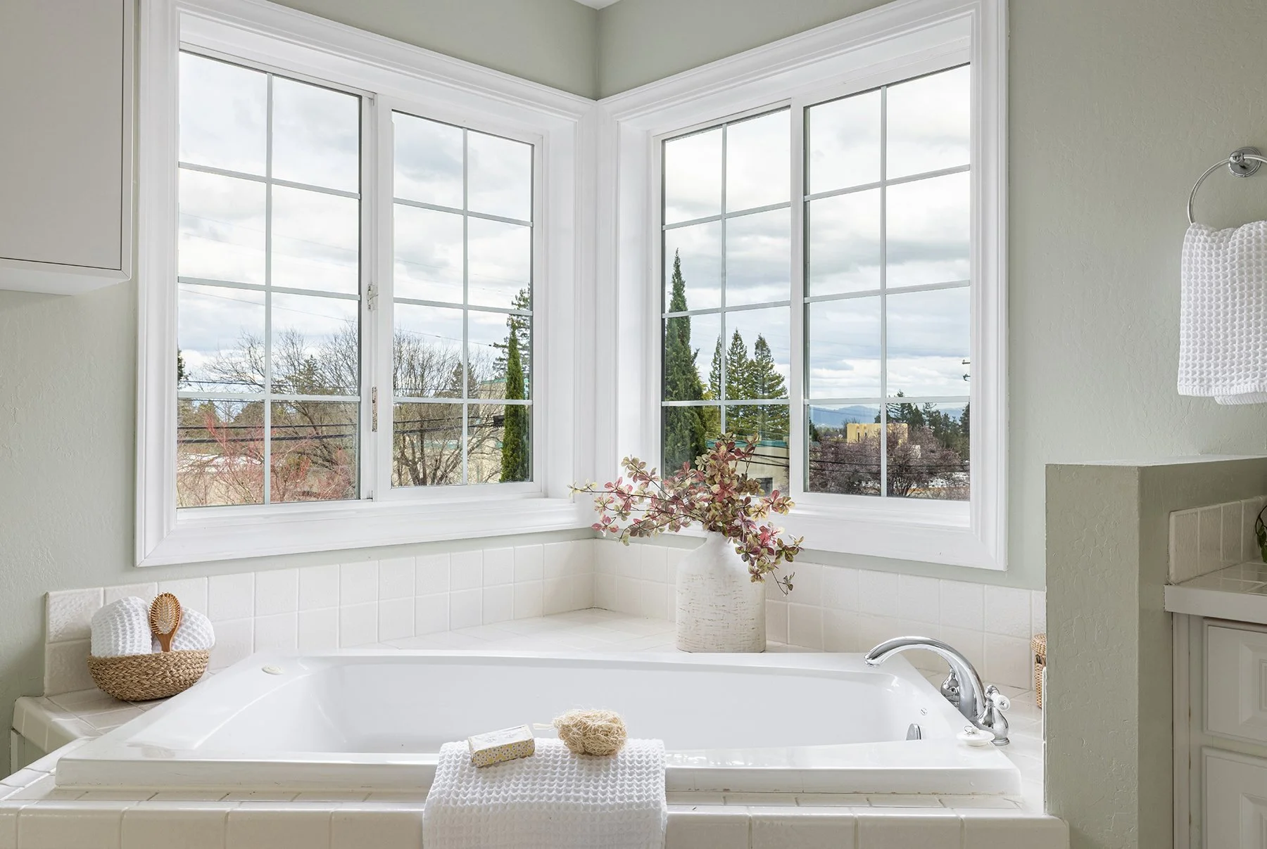 A bathroom bathtub with a large window view of trees and cloudy sky, decorated with a vase of pinkish flowers, a basket of towels and a bath sponge.