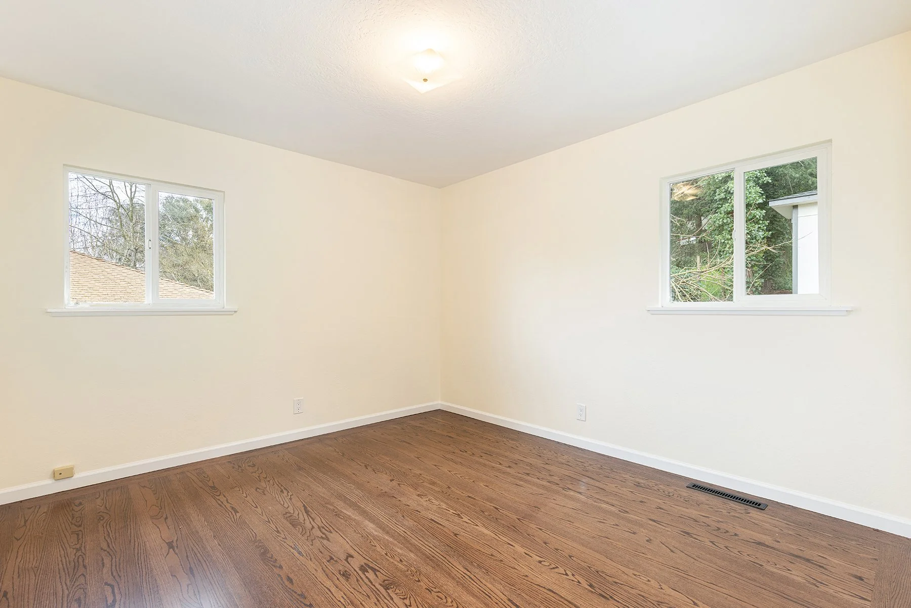 Empty room with beige walls, hardwood floors, two windows, and a ceiling light fixture.