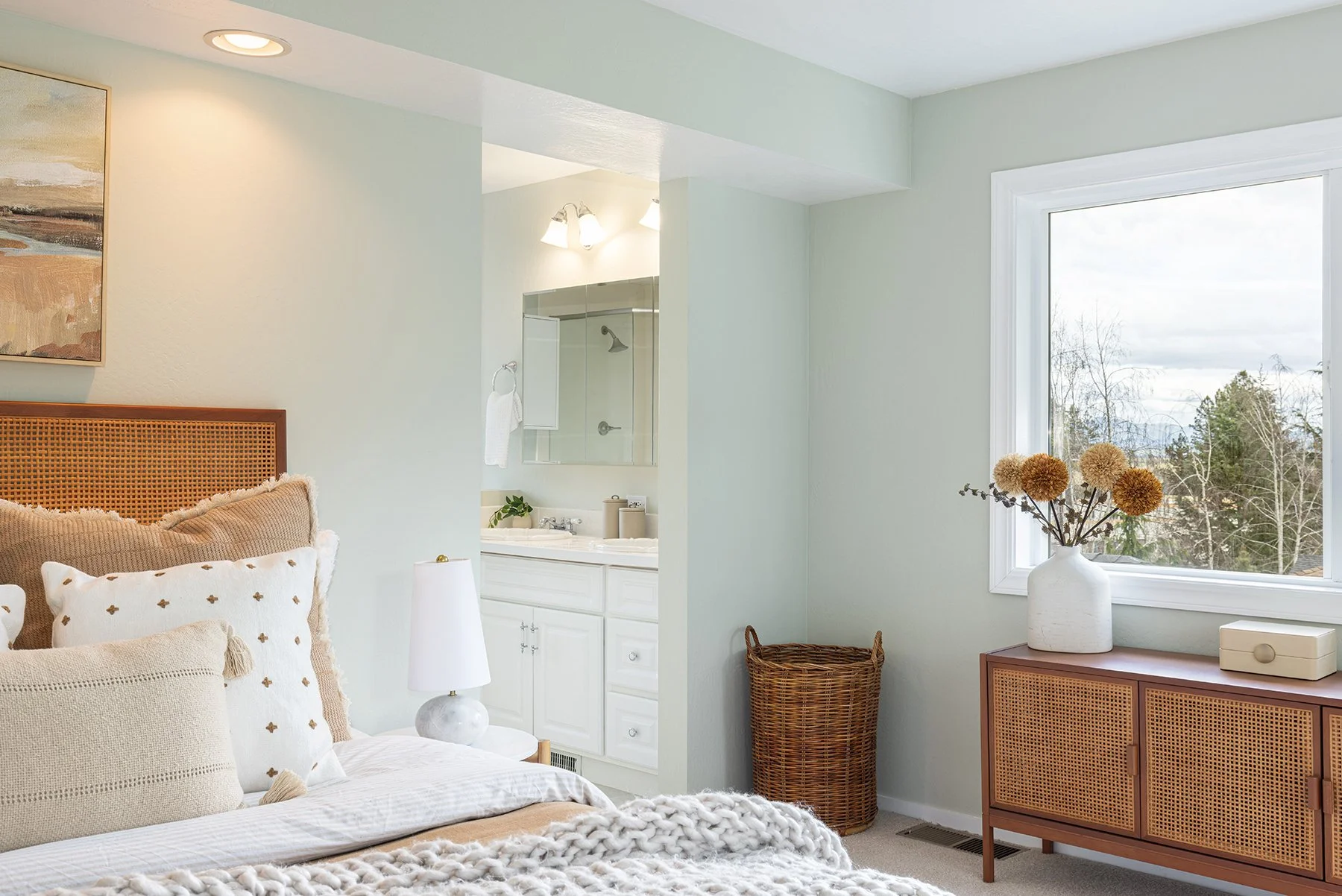 A cozy bedroom with a bed, pillows, nightstand, table lamp, wicker basket, sliding cabinet, and a window with a view of trees and mountains.