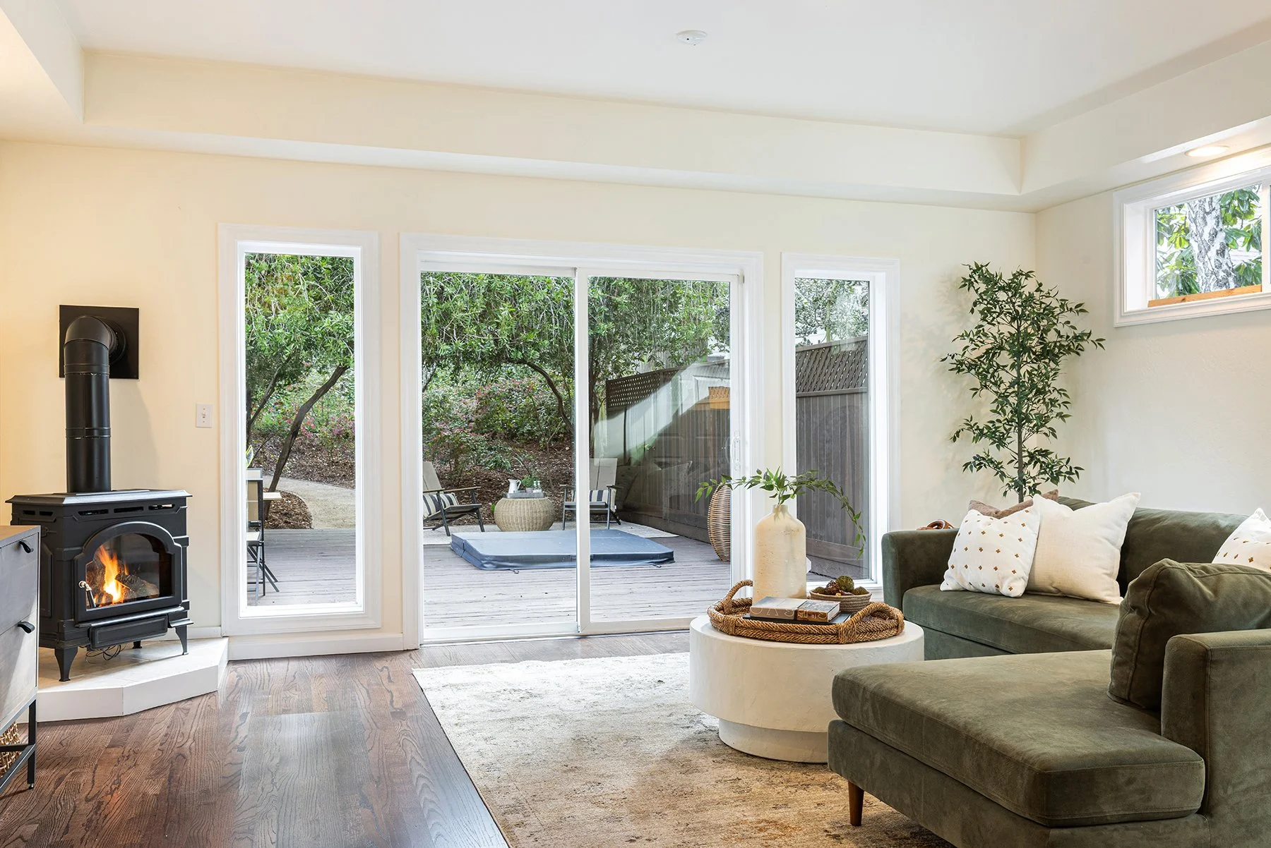Living room with a green sectional sofa, white pillows, wood floor, a white coffee table with a vase and tray, a wood stove with a fire, and large glass sliding doors opening to a backyard patio with outdoor chairs, rug, and trees.