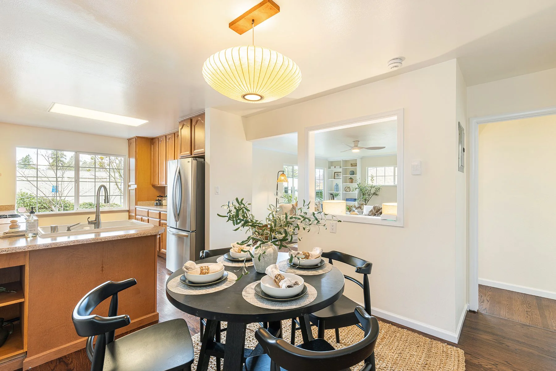 A dining area with a black round table set for four, white bowls with napkins, a large white vase with green foliage, and woven placemats on a natural fiber rug. Adjacent to the kitchen, which has wooden cabinets, a stainless steel refrigerator, and 