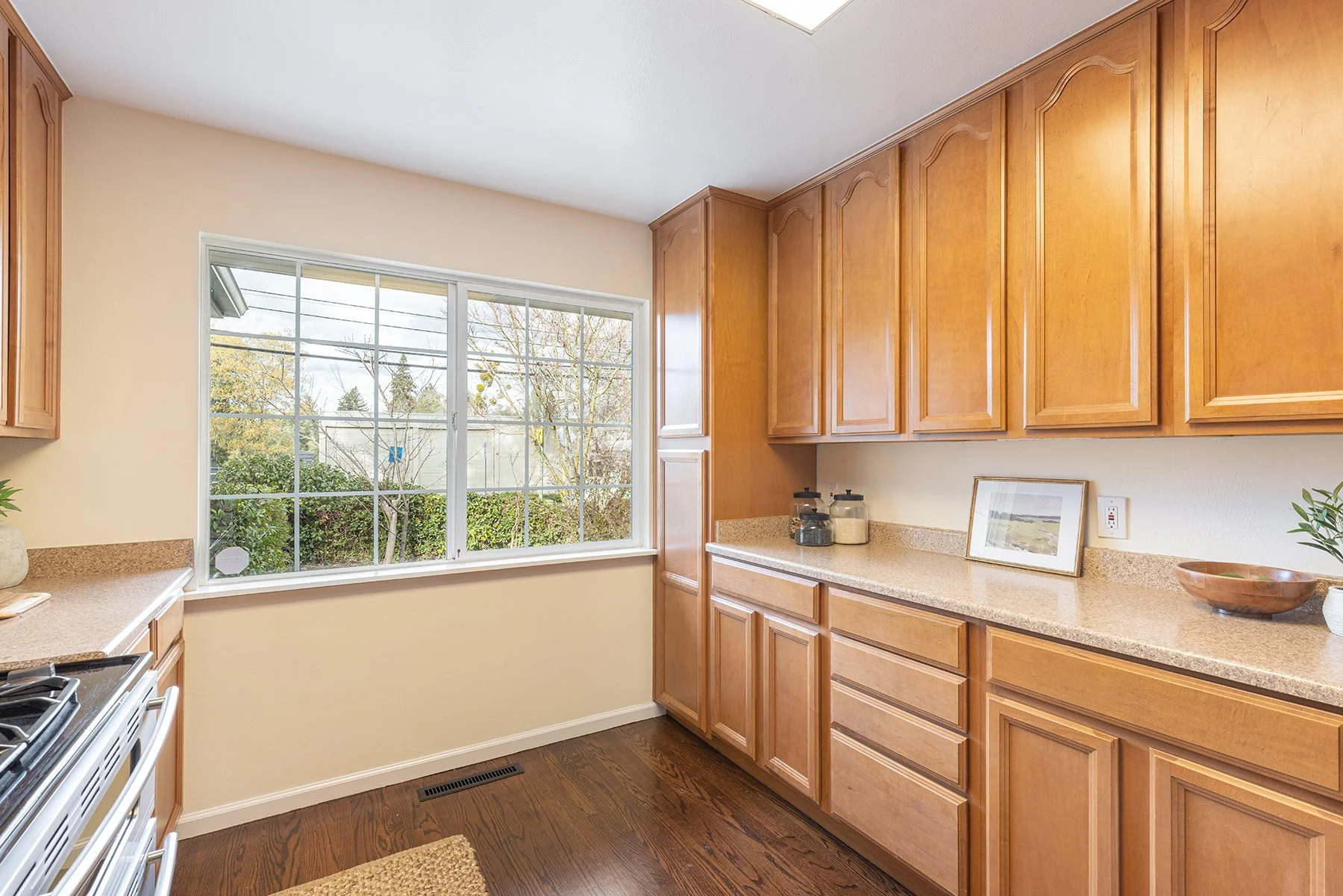 Kitchen with wooden cabinets, beige countertops, a window view of trees, and a framed picture on the counter.
