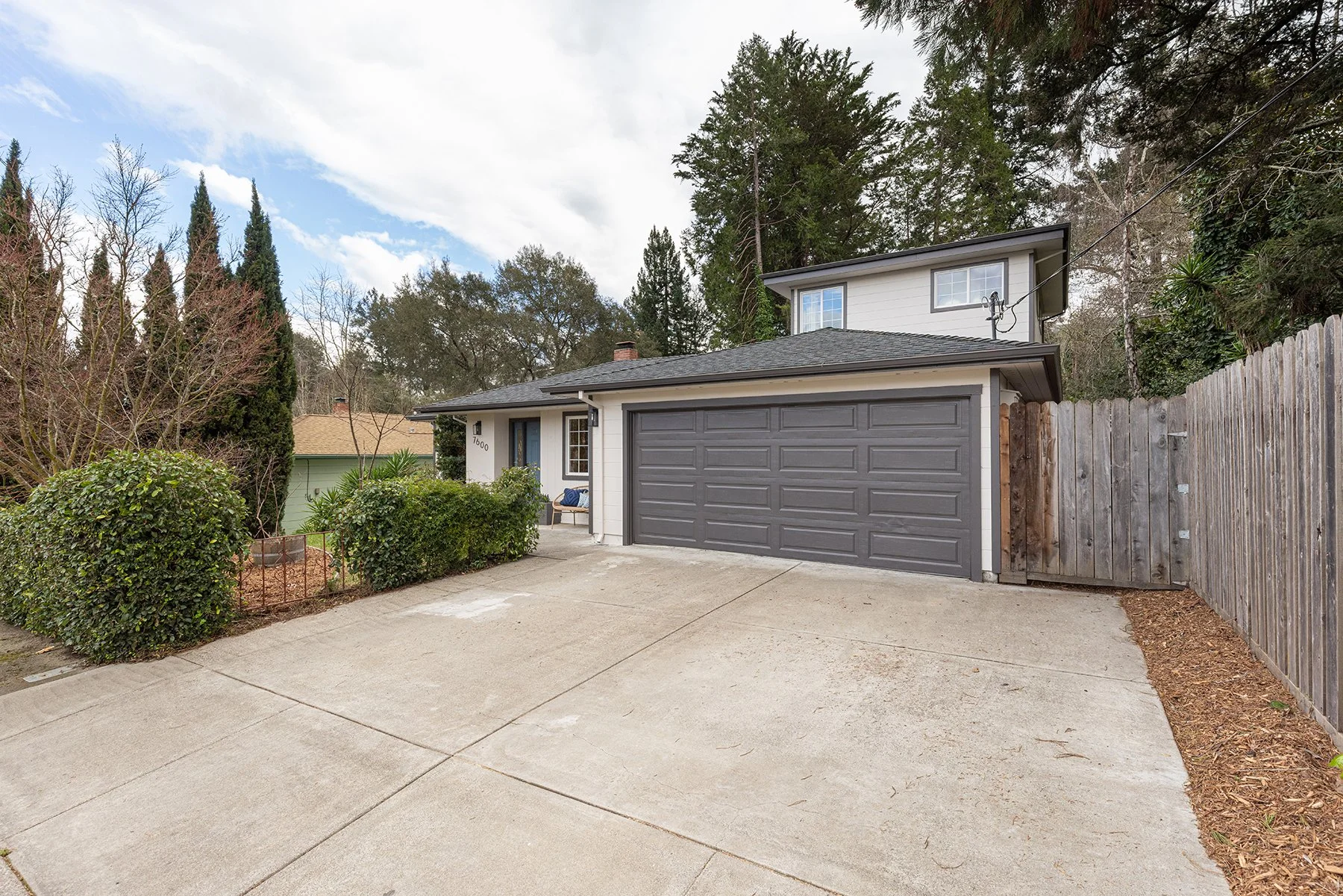 Modern two-story house with a gray garage door, driveway, and wooden fence, surrounded by trees and shrubs under a partly cloudy sky.