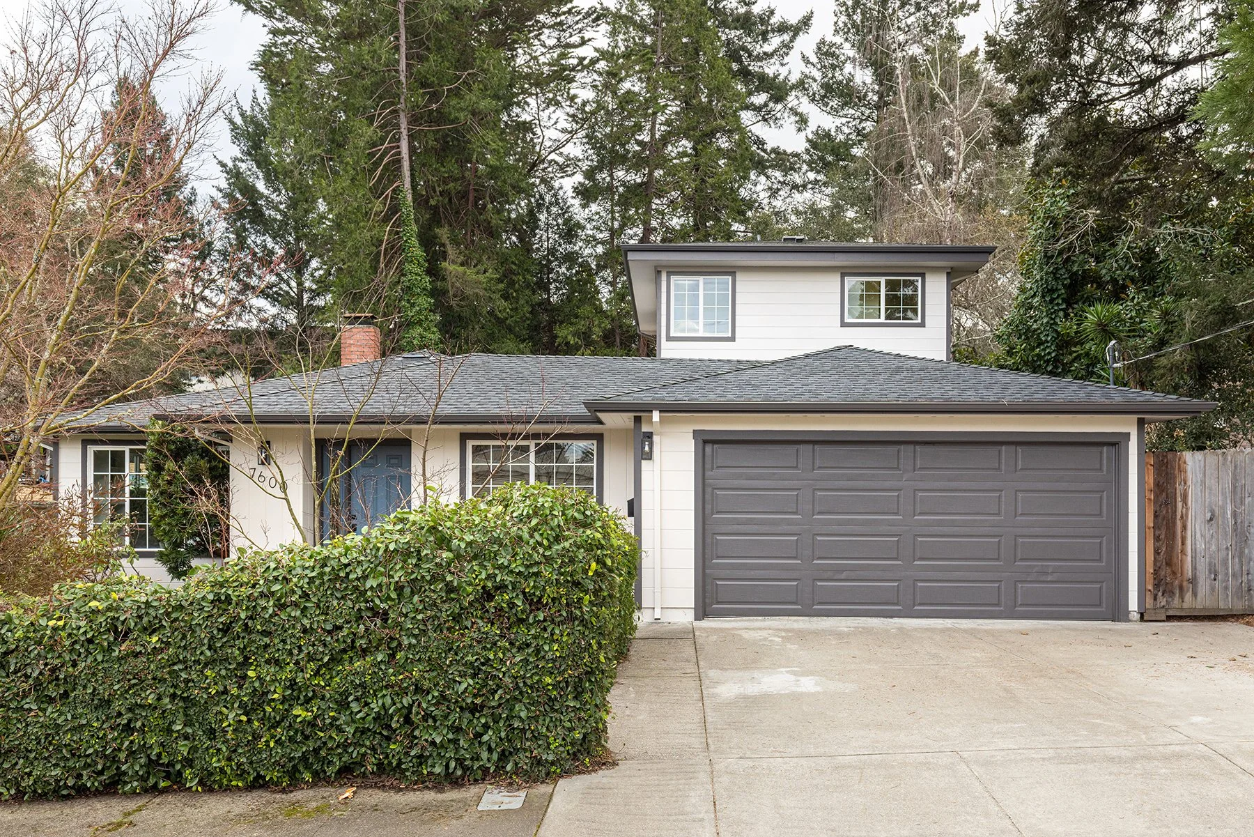 Front view of a modern two-story house with gray garage door, white siding, and black window trim, partially obscured by a green hedge.