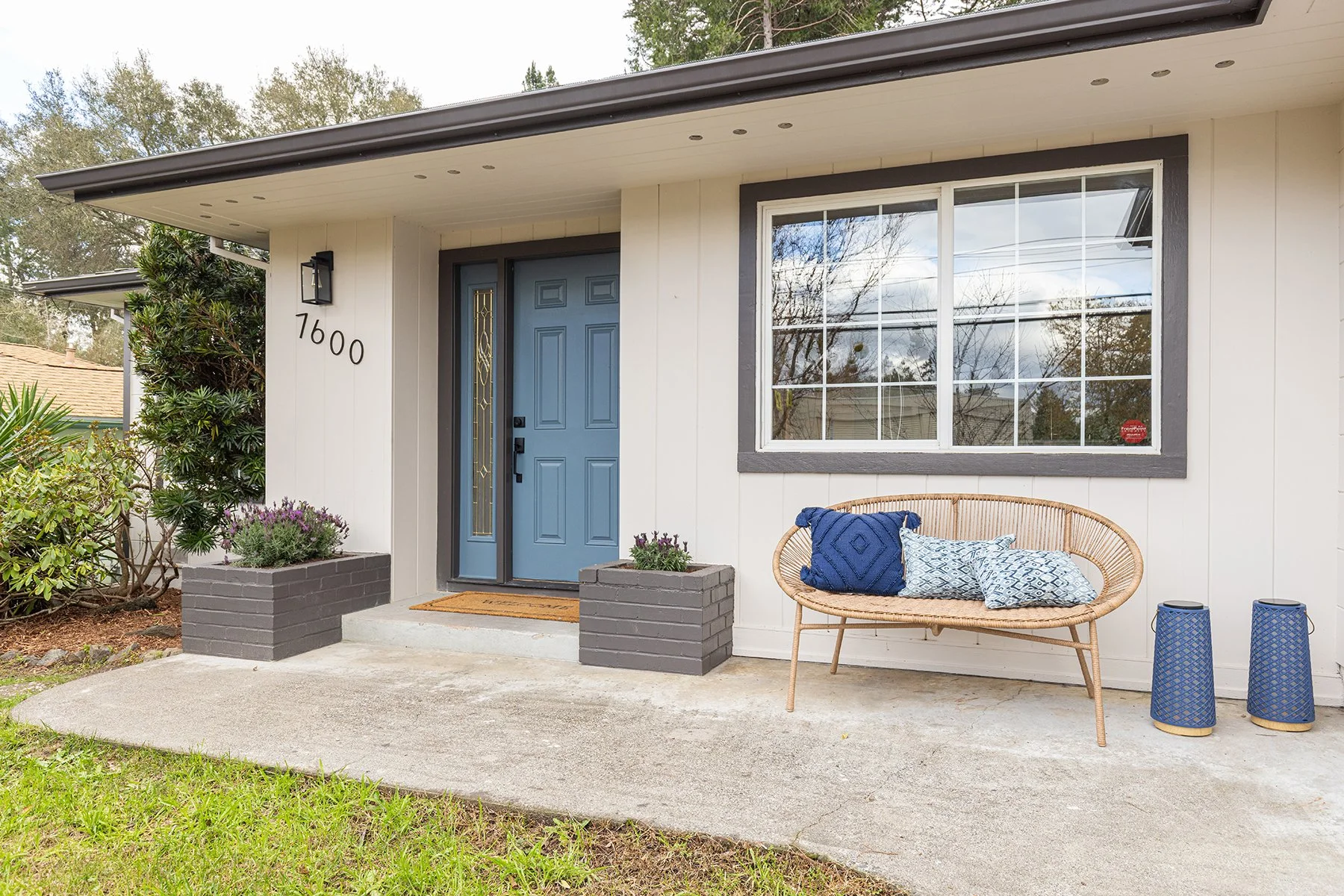 Front porch of a house with a pale blue door, large window with gray trim, a woven bench with blue and patterned cushions, and two blue lantern-style planters. The house number 7600 is on the wall, with greenery on the side and plants in gray planter