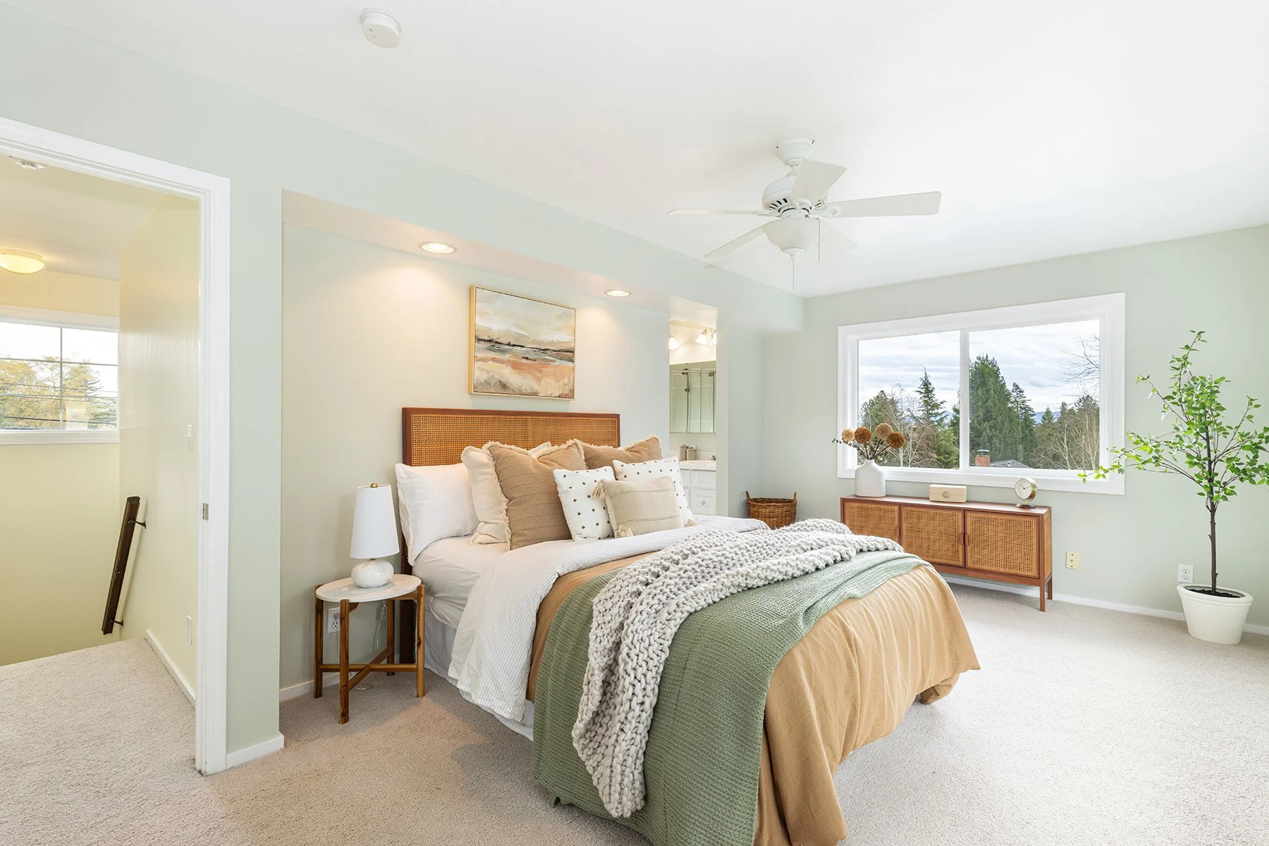 Bright bedroom with a large window, bed with beige and white linens, green and gray blankets, and multiple pillows. There is a white nightstand with a lamp, a wooden cabinet with decor, and potted plants.