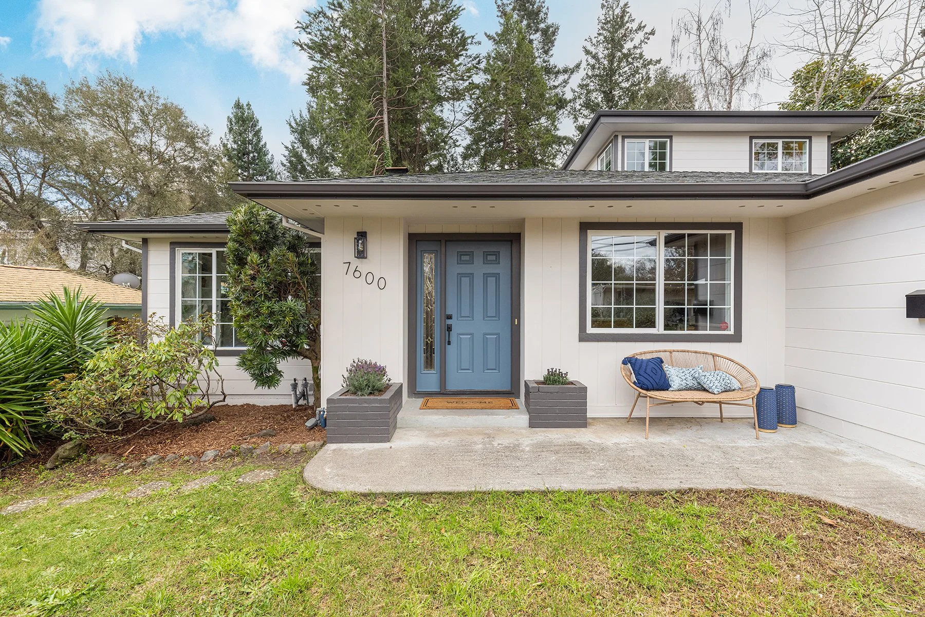 Front entrance of a modern house with a blue door, large window, and outdoor seating, surrounded by greenery.