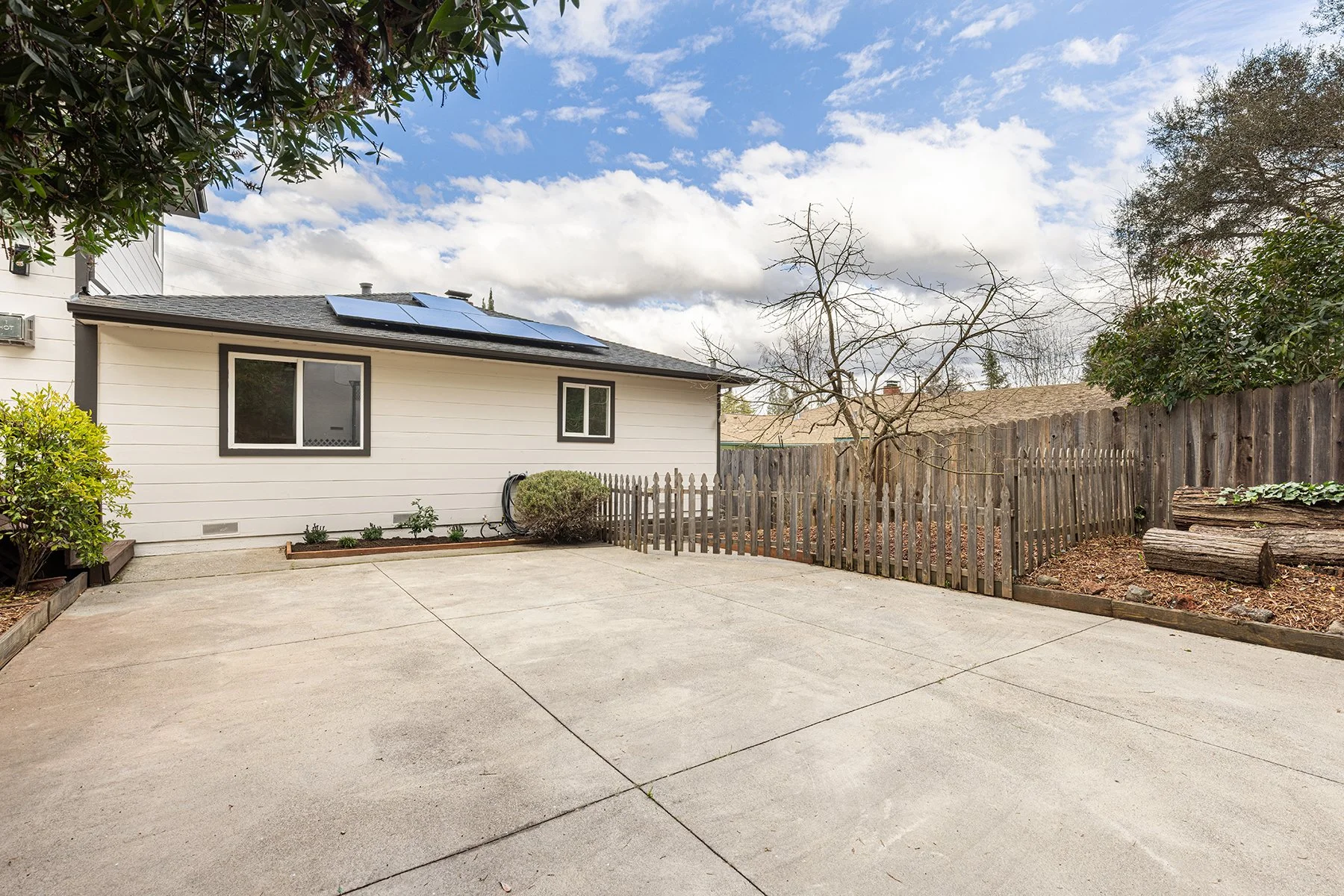 Backyard with a concrete patio, a white house with solar panels, a small garden bed, a sagging fence, a leafless tree, and logs on a hillside, under a partly cloudy sky.