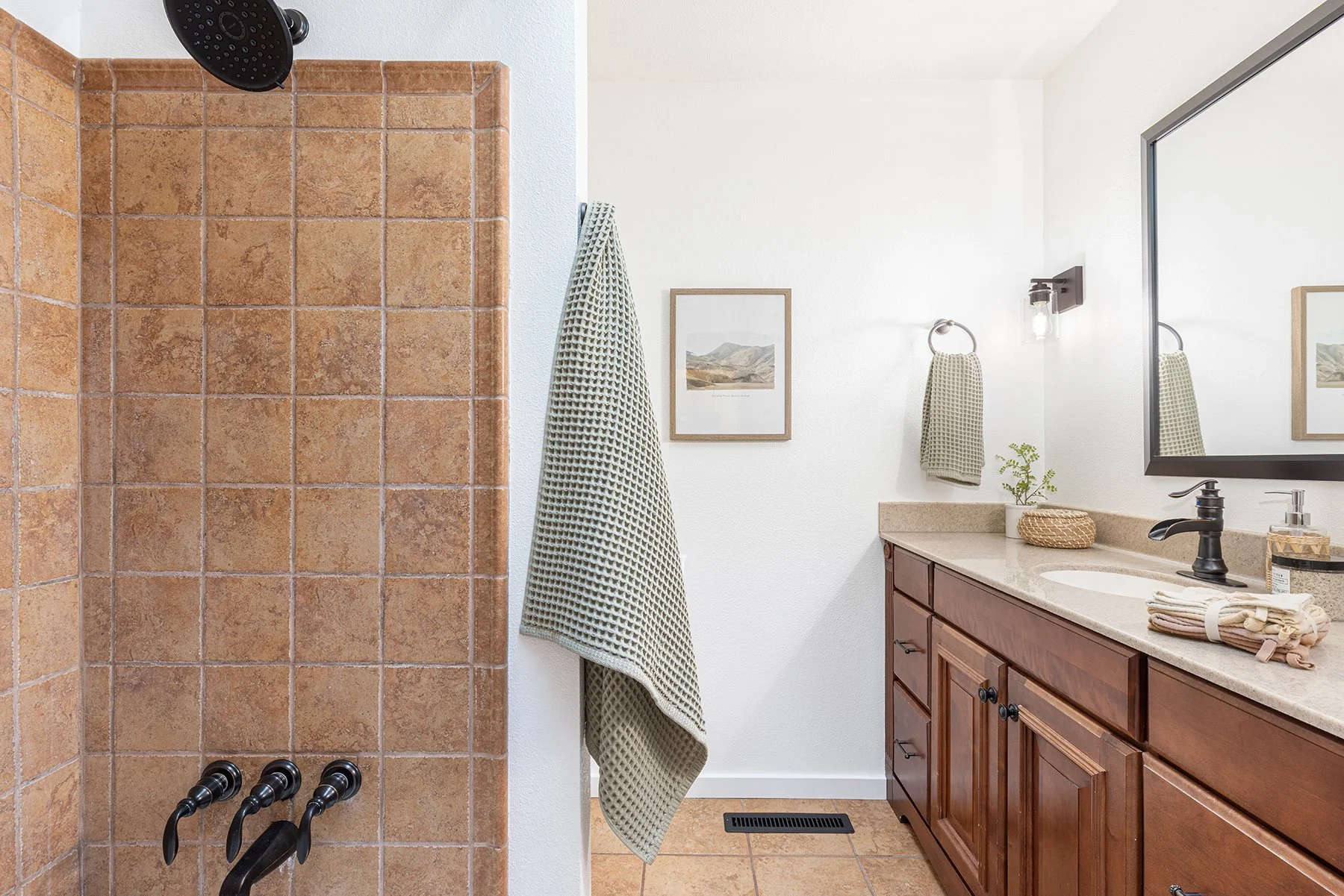 Bathroom with a walk-in shower with brown tiles, wooden vanity with a granite countertop, a large mirror, a framed picture on the wall, a towel, a soap dispenser, and a small plant.