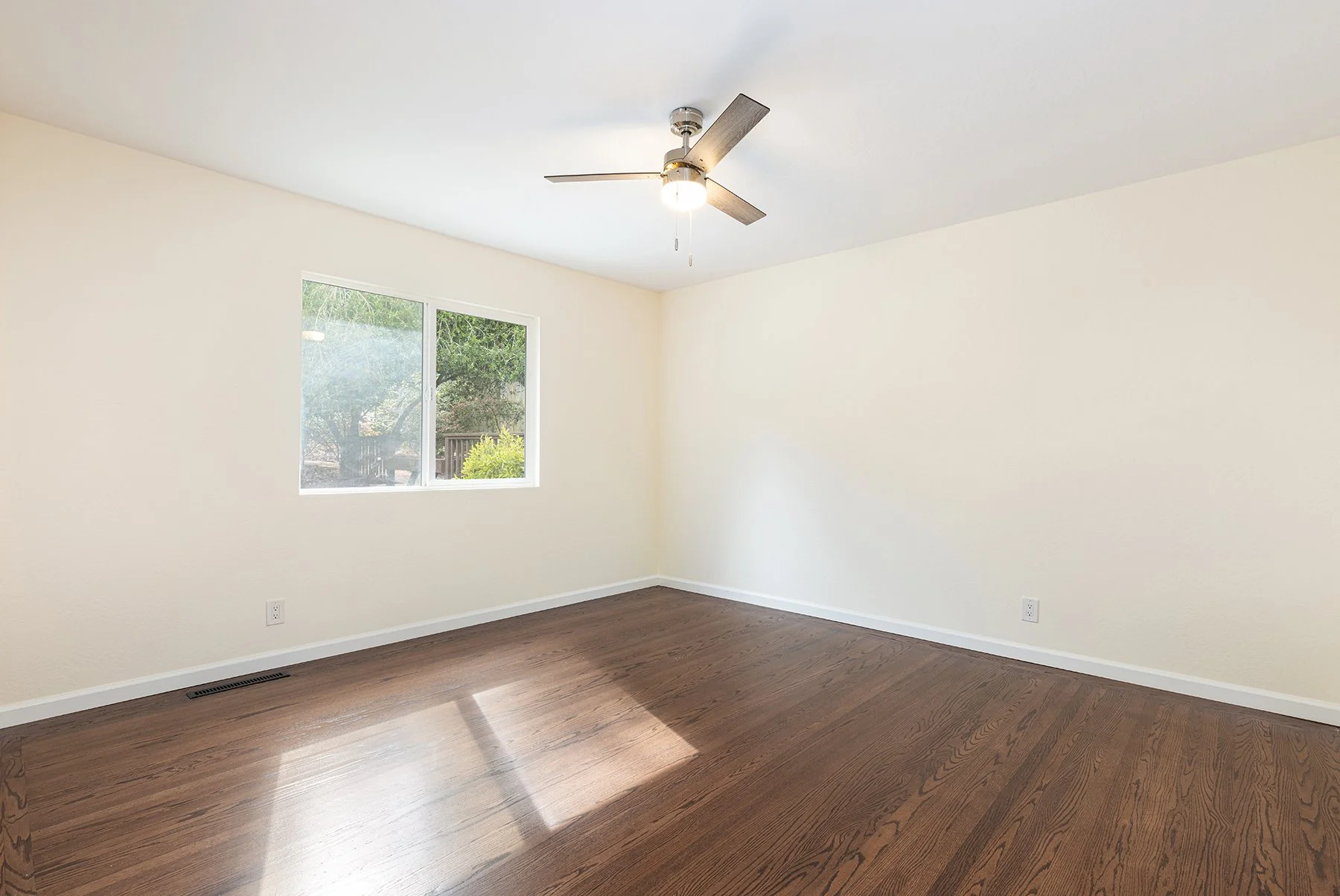 Empty room with white walls, hardwood flooring, a window showing outdoor trees and greenery, a ceiling fan with a light fixture, and electrical outlets.