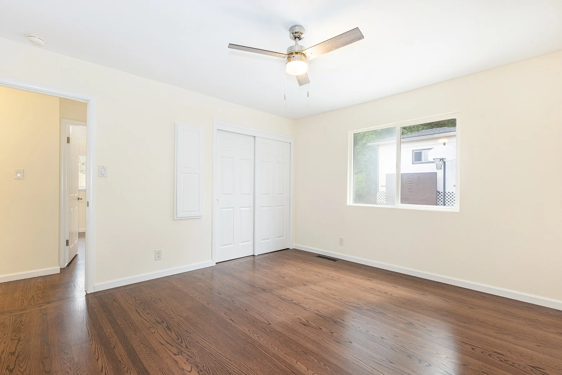 Empty living room with hardwood floors, white walls, a ceiling fan, and a window showing the backyard with a basketball hoop.