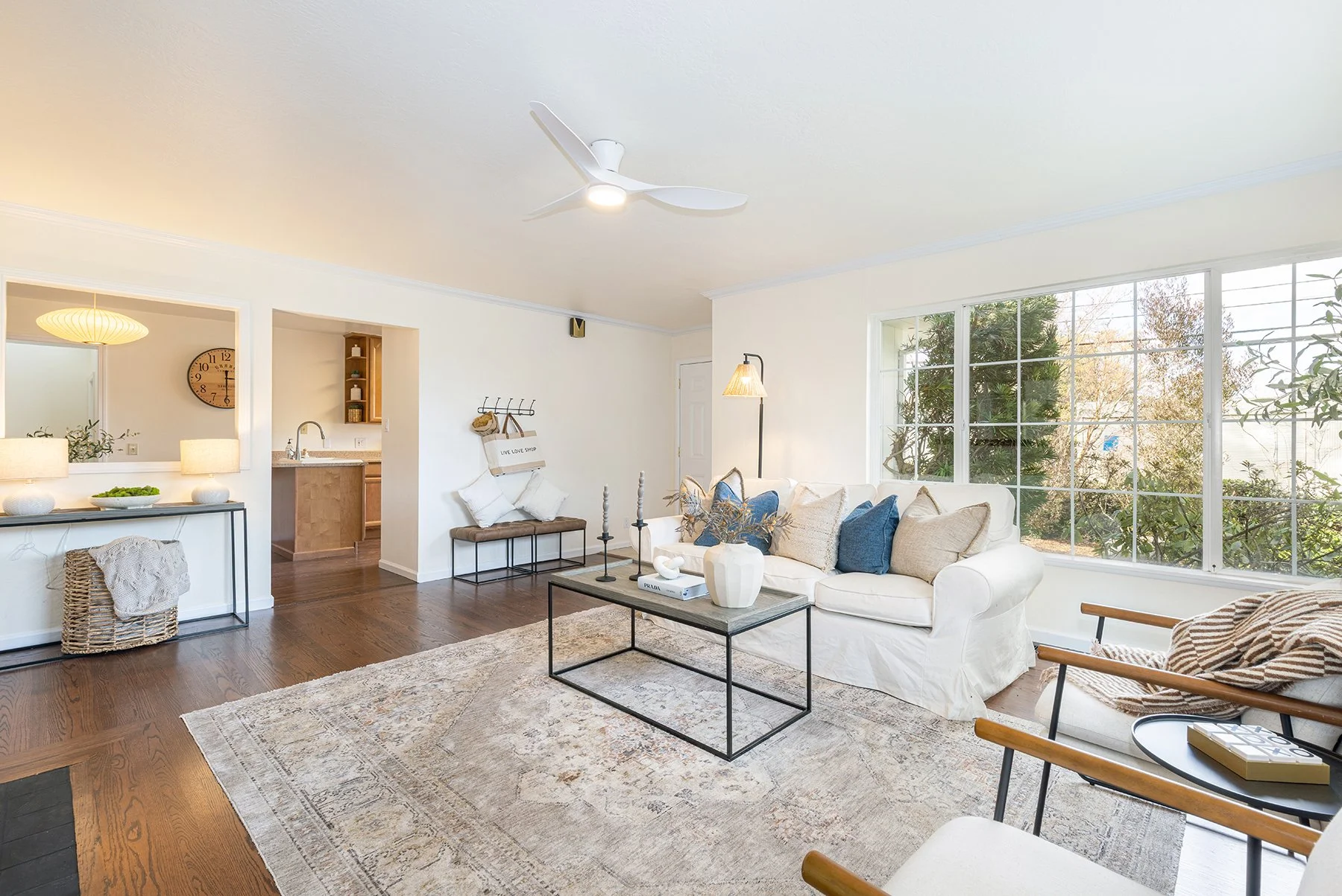 Bright living room with large window, white sofa with blue and beige pillows, wooden chairs with striped blanket, black coffee table with white vase, and large patterned area rug.