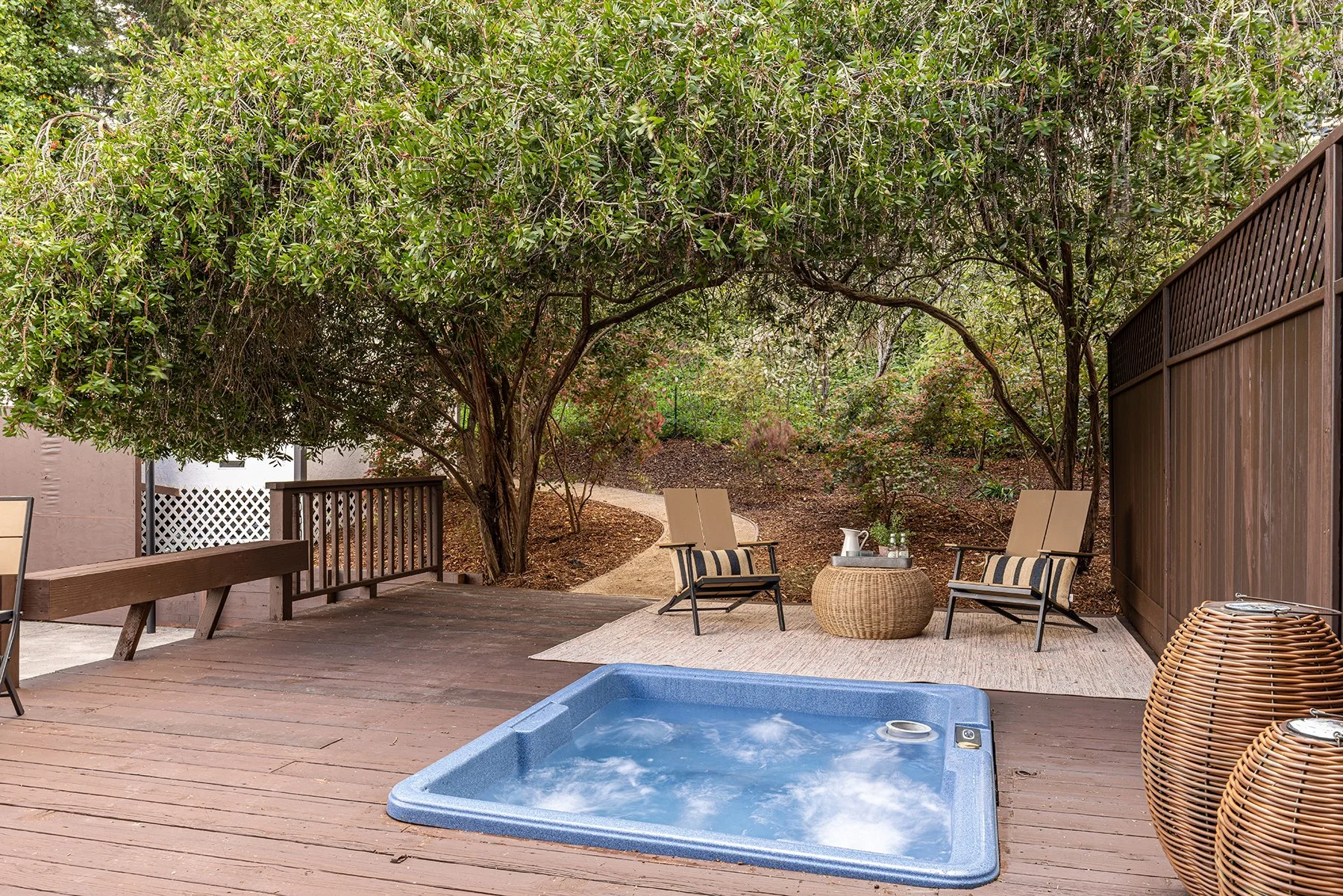 A backyard wooden deck with a blue hot tub, outdoor chairs, a wicker side table with drinks, a rug, and a large leafy tree providing shade over the sitting area.