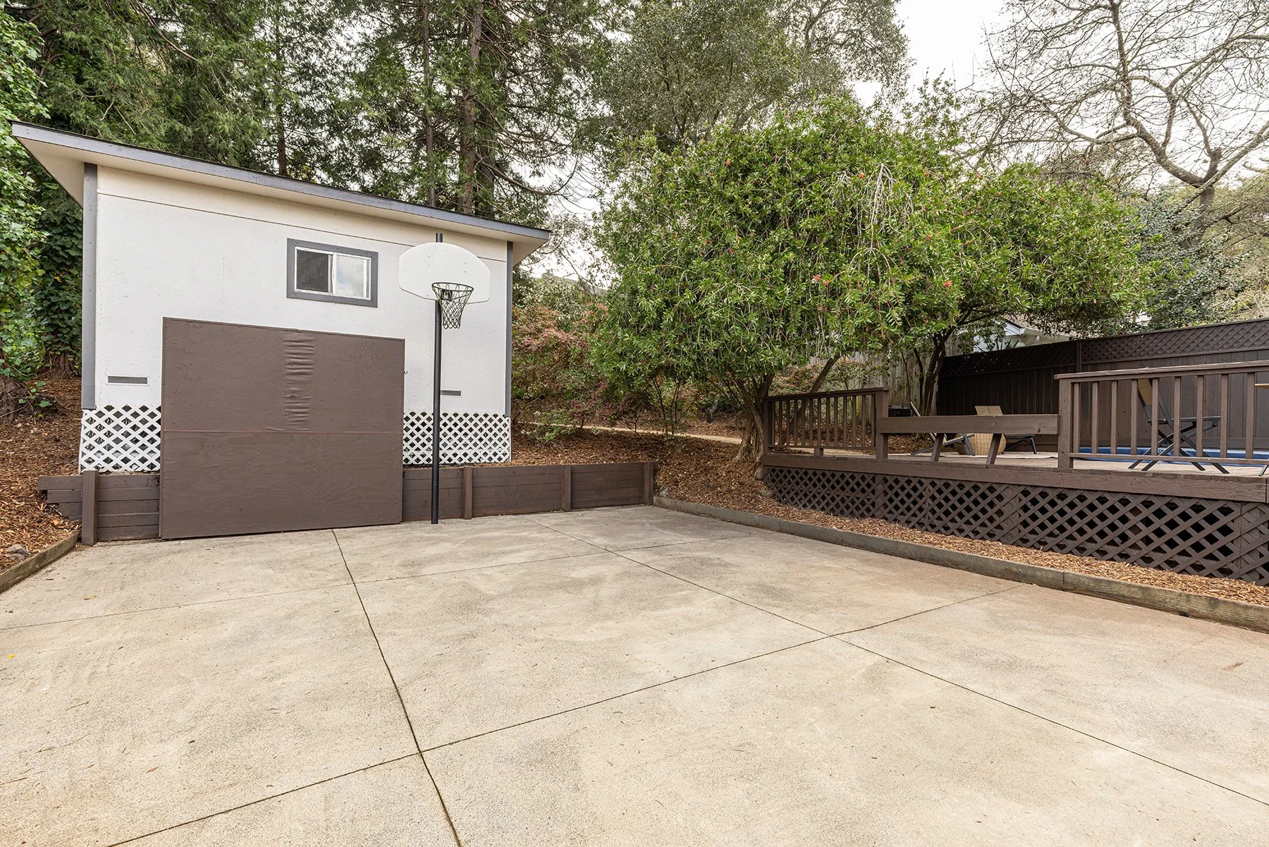 Empty backyard with a concrete patio, a baseball hoop, a small shed, a wooden deck with outdoor furniture, surrounded by trees and a fence.