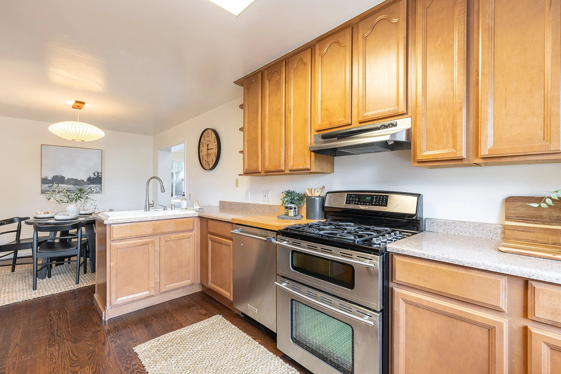 Kitchen with wooden cabinets, stainless steel oven, and gas stove, open to a dining area with a black table and chairs, and wall art.