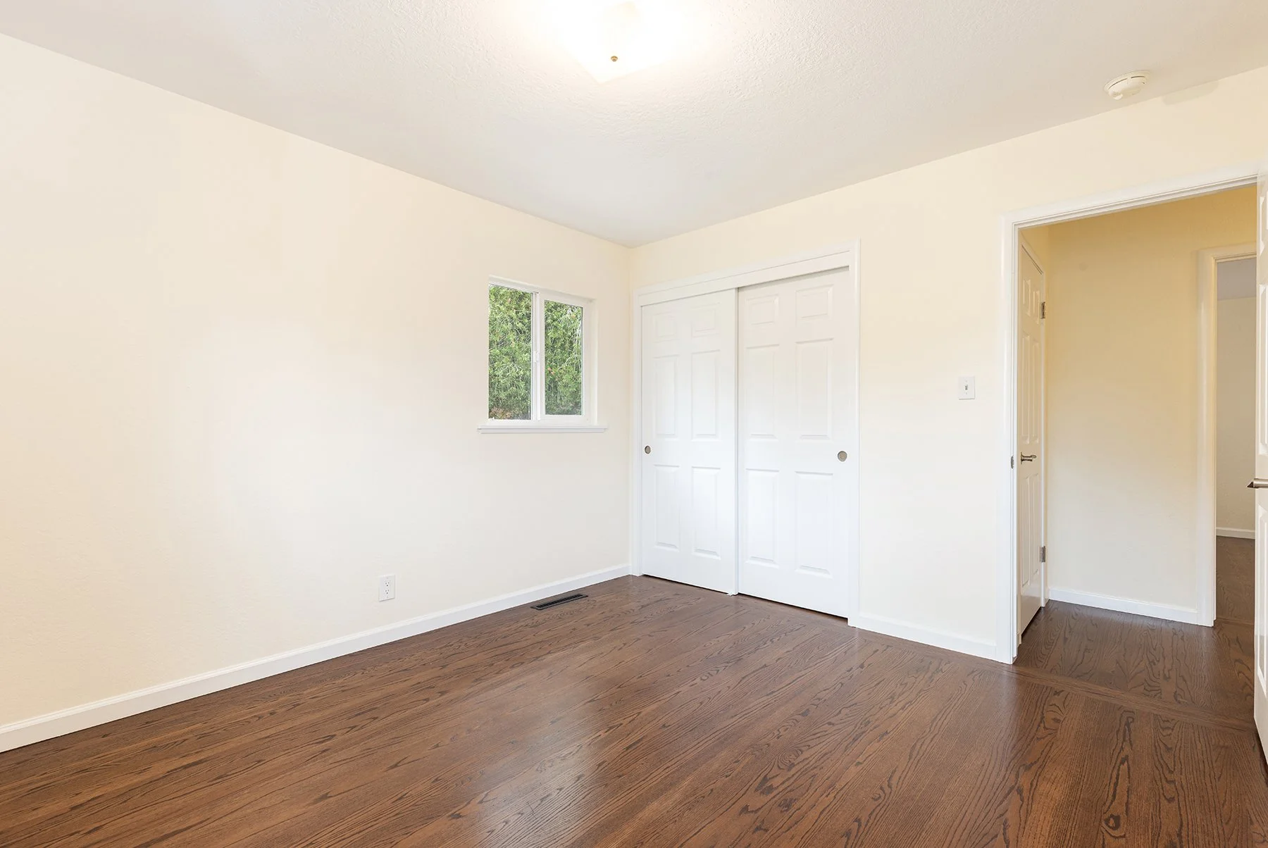Empty bedroom with white walls, hardwood floor, small window, and a closet with sliding doors.