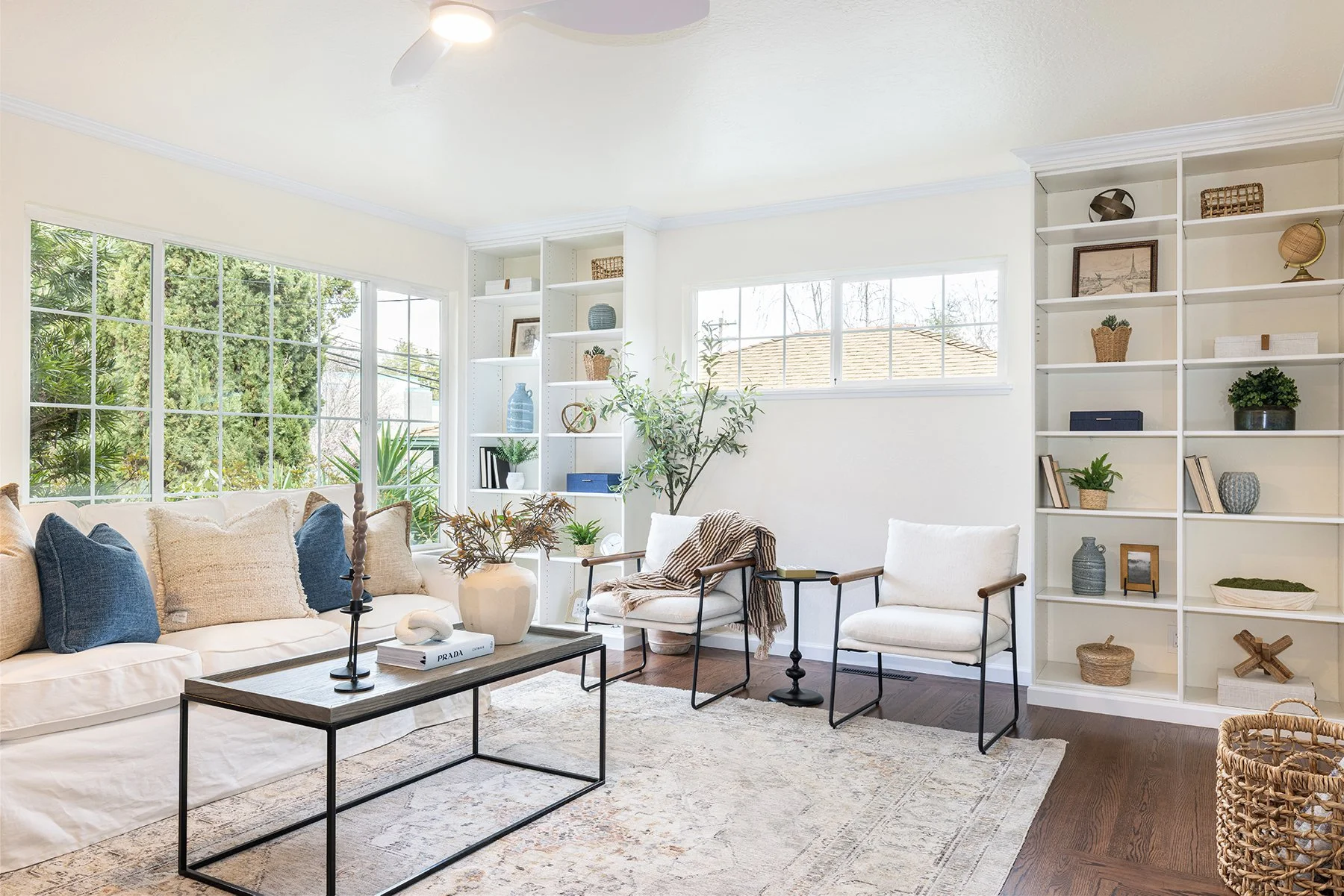 Bright living room with white sofa, beige and blue pillows, and black coffee table. Large windows with greenery outside and built-in white shelves with decorative items and books. Two white chairs with wooden arms and a throw blanket. Light colored a