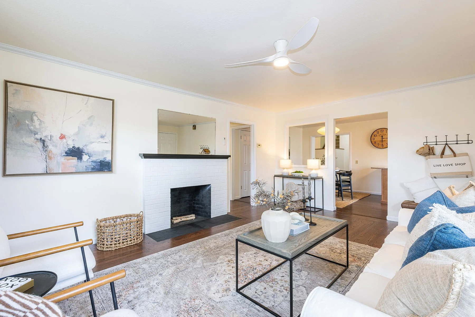 Living room with white walls, hardwood flooring, a white brick fireplace, modern art on the wall, a ceiling fan, and cozy furniture including a sofa with blue pillows and a wooden armchair.