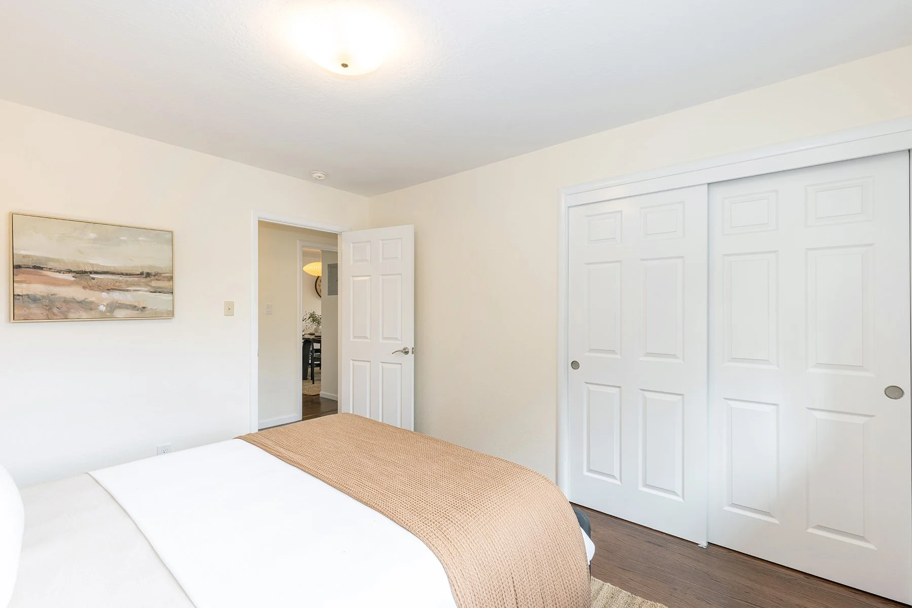A bedroom with white walls, a ceiling light, a framed landscape painting, a partially visible bed with white sheets and a tan blanket, a closet with white sliding doors, an open door leading to a hallway, and dark wood flooring.