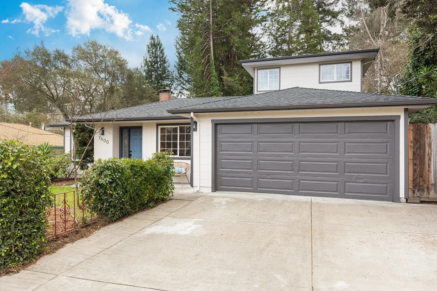 A two-story house with a gray garage door, a blue front door, and white siding, surrounded by trees and bushes, with a concrete driveway and sidewalk.