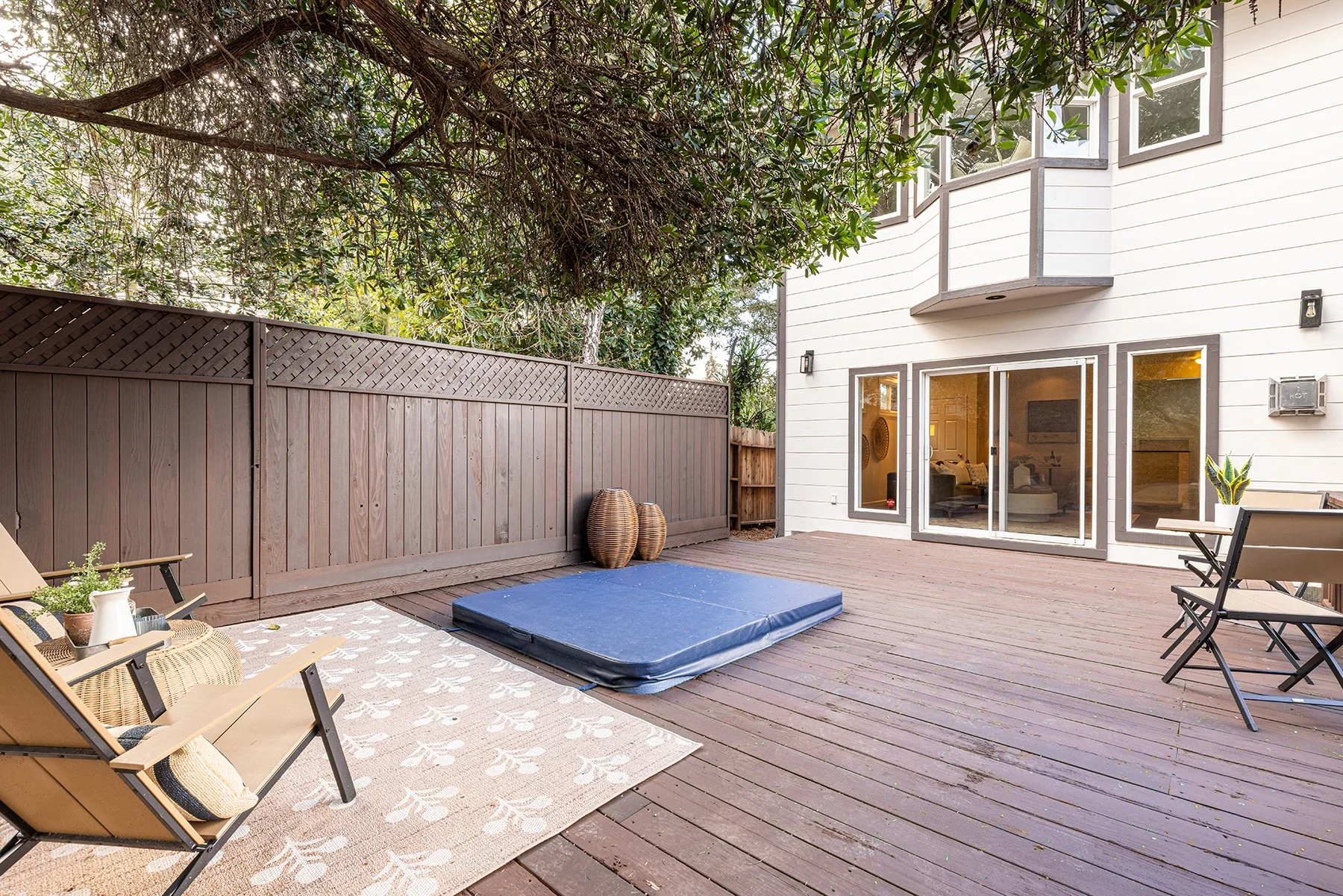A backyard patio with wooden decking, enclosed by a brown fence, featuring two chairs with cushions, a patterned outdoor rug, a blue gymnastics mat, and decorative vases, adjacent to a white house with large sliding glass doors and windows.