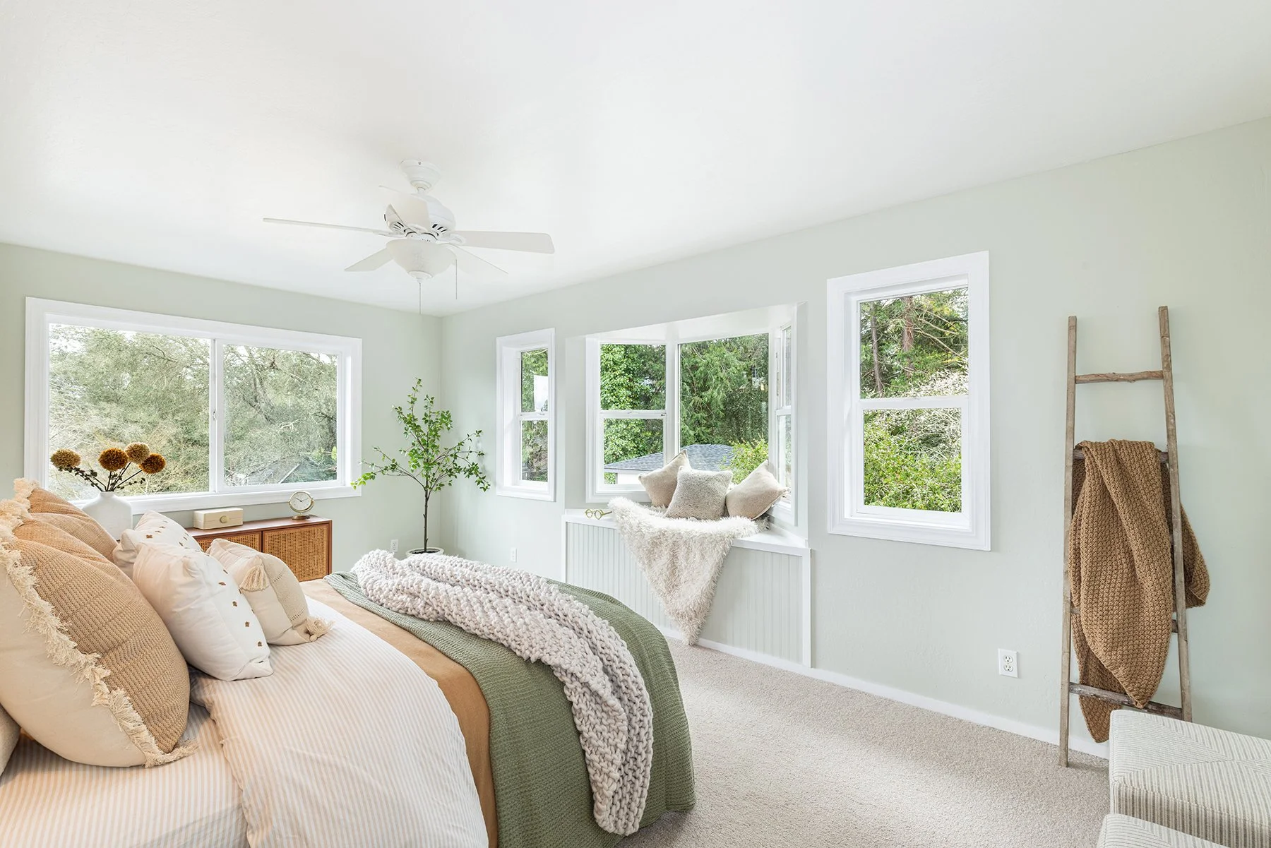 Bright bedroom with large window, featuring a bed with beige and cream pillows, a throw blanket, a small plant, a ladder with blankets, and a window seat with pillows.