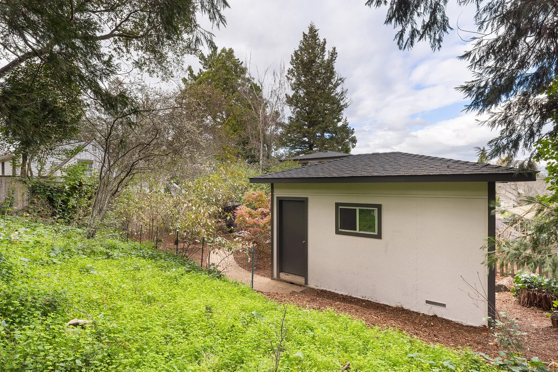Backyard with green grass, trees, and a small white building with black trim and a black door.