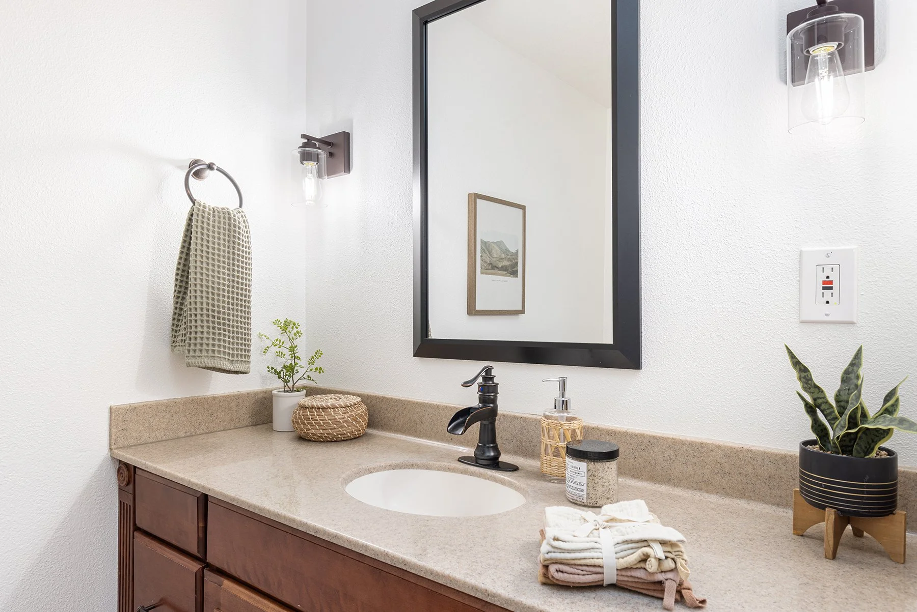Bathroom vanity with a beige countertop, black faucet, mirror, wall-mounted lights, plants, and folded towels.