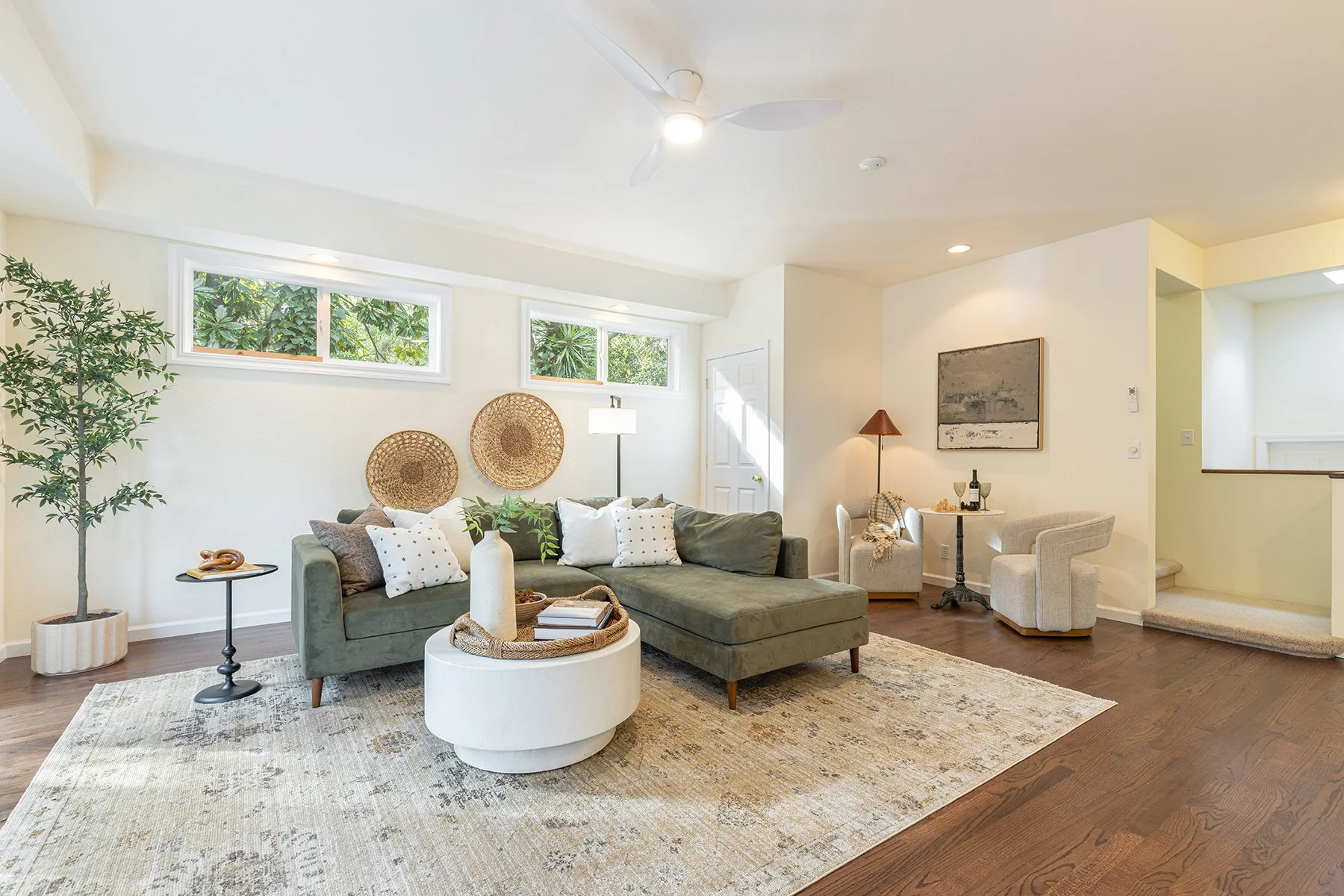 Bright living room with large L-shaped green sofa, decorative pillows, and a white round coffee table, surrounded by natural light from high windows, with side chairs, a painting, and wooden flooring.