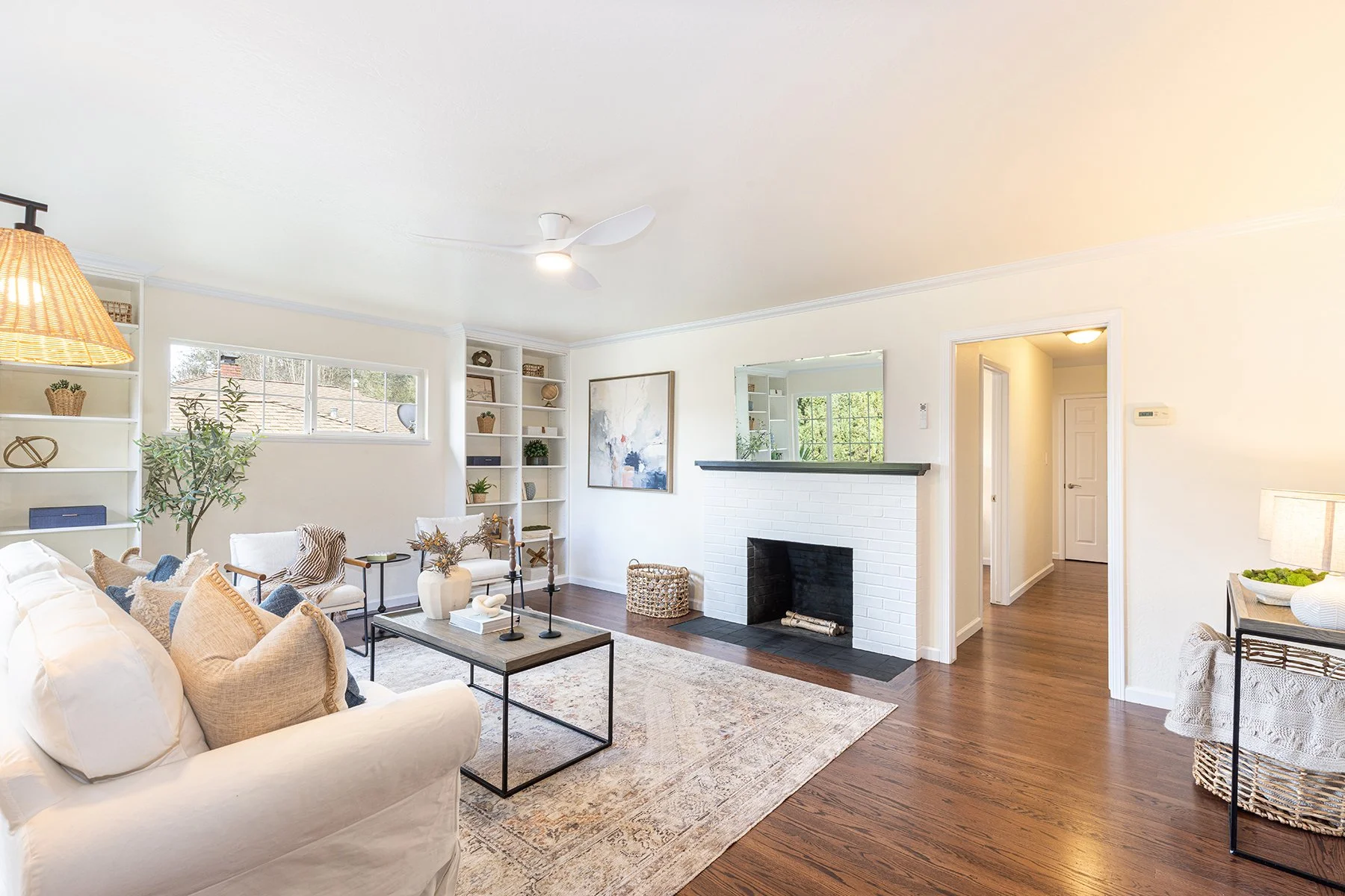 Bright living room with white sofa, armchairs, a coffee table, built-in shelves, a fireplace, and a window, decorated with plants, artwork, and home accessories.