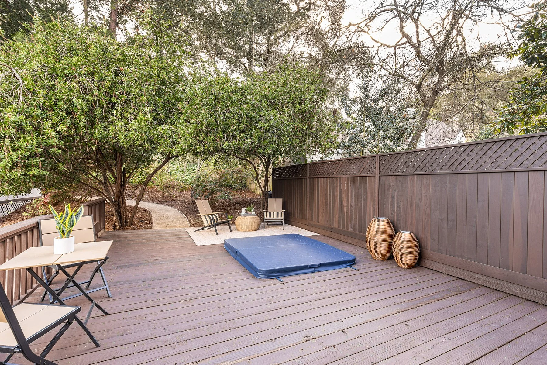 Backyard patio with wooden deck, inflatable bounce house, outdoor chairs, table with plant, and decorative vases, surrounded by trees and a fence.