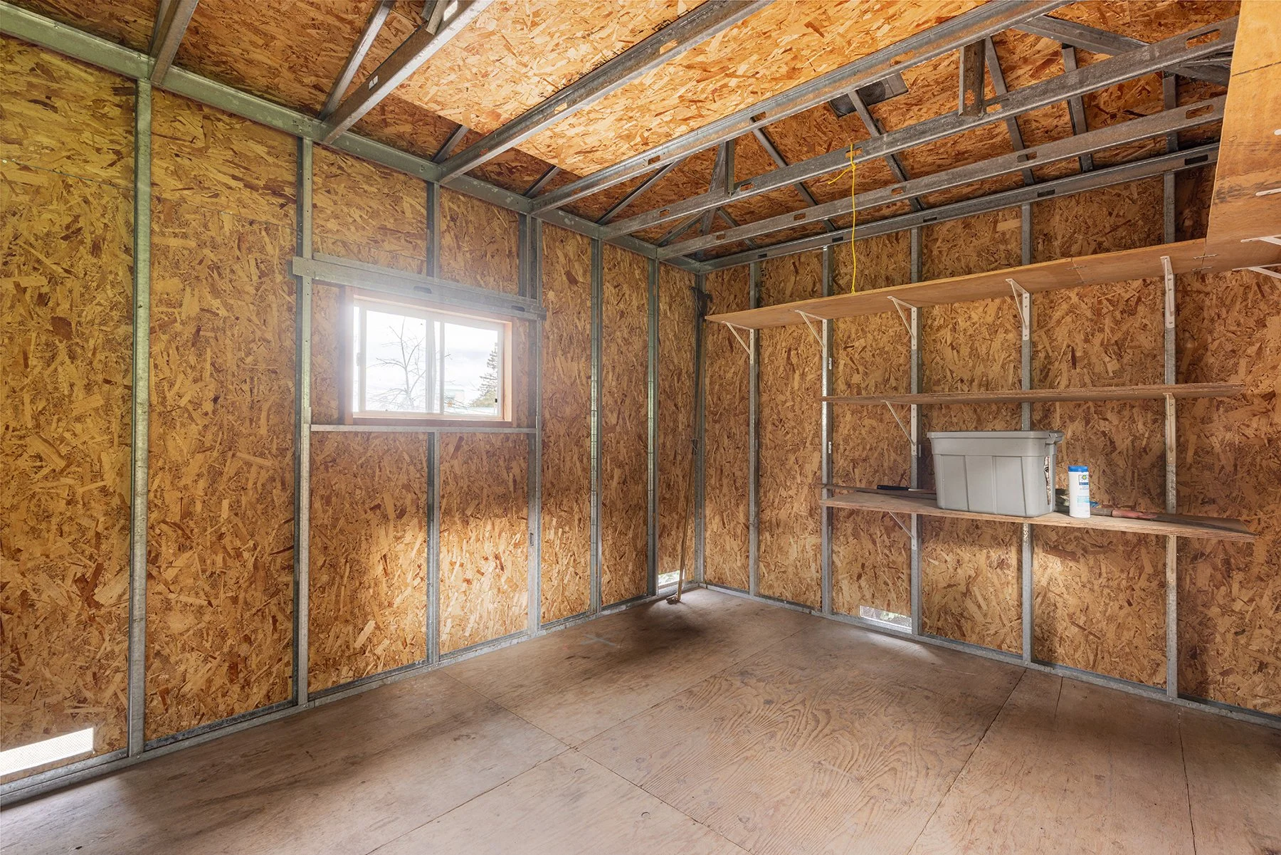 Interior of a partially constructed shed with plywood walls, metal framing, a window, and suspended wooden shelves, with construction materials on one shelf.