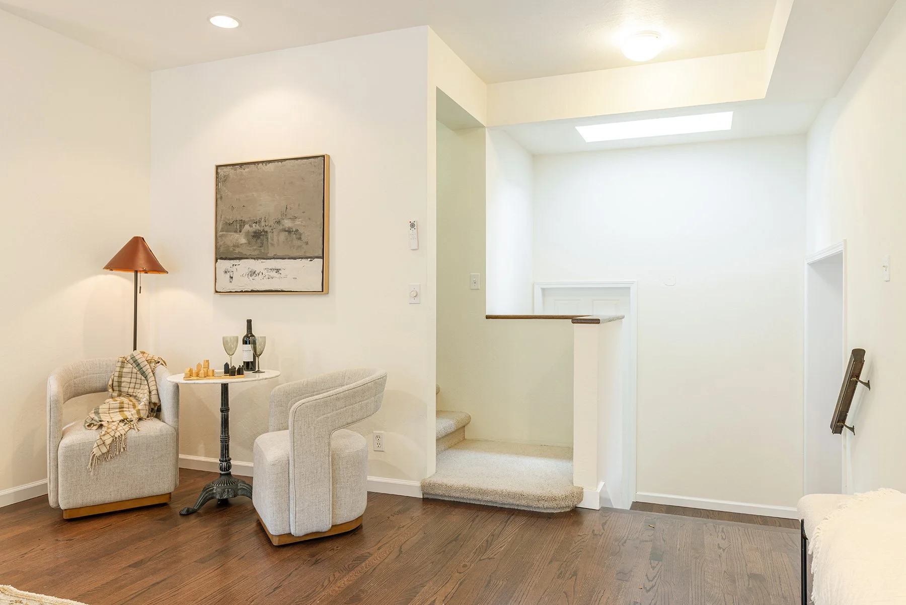 Living room with white walls and a hardwood floor. There is a small seating area with two light-colored armchairs, a small round table with wine and glasses, and a floor lamp. A painting hangs on the wall. A staircase with carpeted steps is in the ba