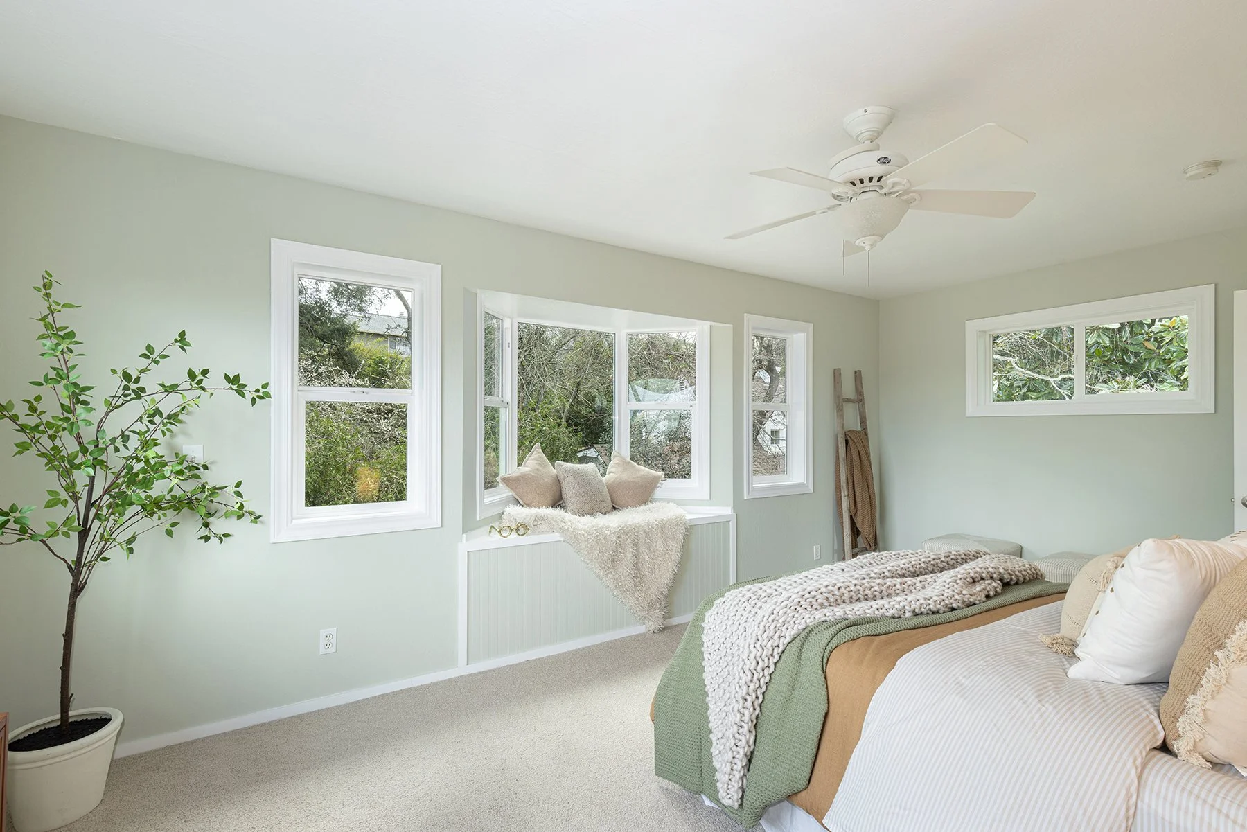 A bedroom with a large bed, multiple pillows, a blanket, and a quilt. There is a potted plant in the corner and a window seat with cushions and a faux fur throw. The room has multiple windows, a ceiling fan, and light-colored walls.