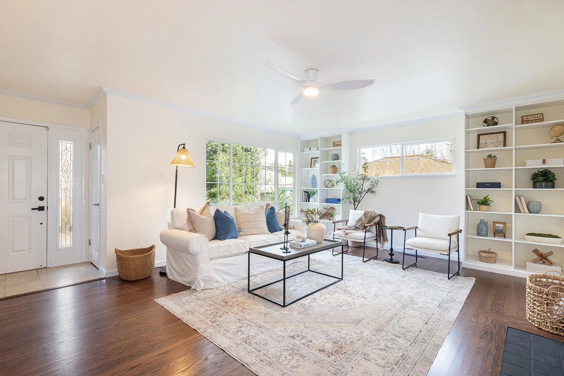 Bright living room with white walls, large windows, a white sofa with throw pillows, two white armchairs, a black coffee table, a modern floor lamp, built-in white bookshelves with decor, and a beige area rug on dark hardwood floors.
