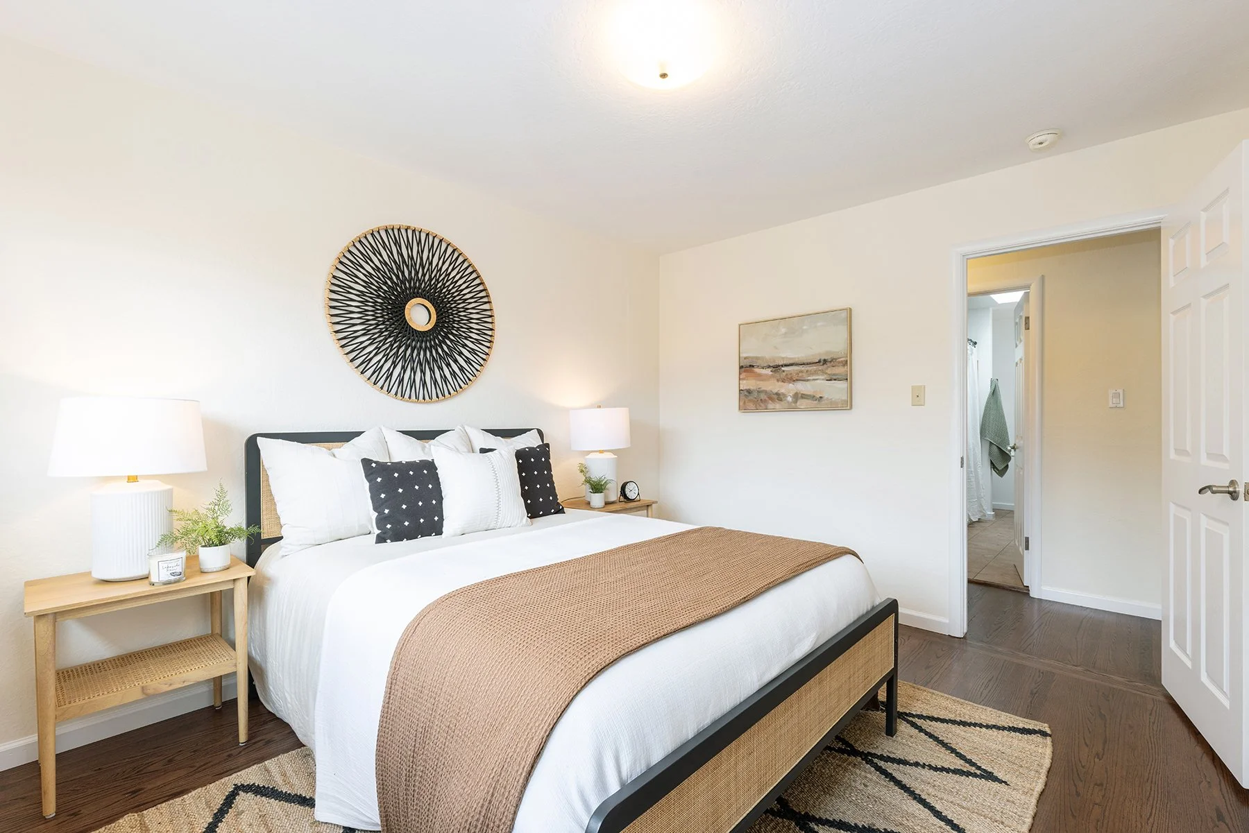 Modern bedroom with white walls, a large bed with white and black pillows, a beige throw blanket, and two wooden nightstands with lamps. Decor includes a circular woven wall art above the bed and a landscape painting on the wall. There is a doorway l