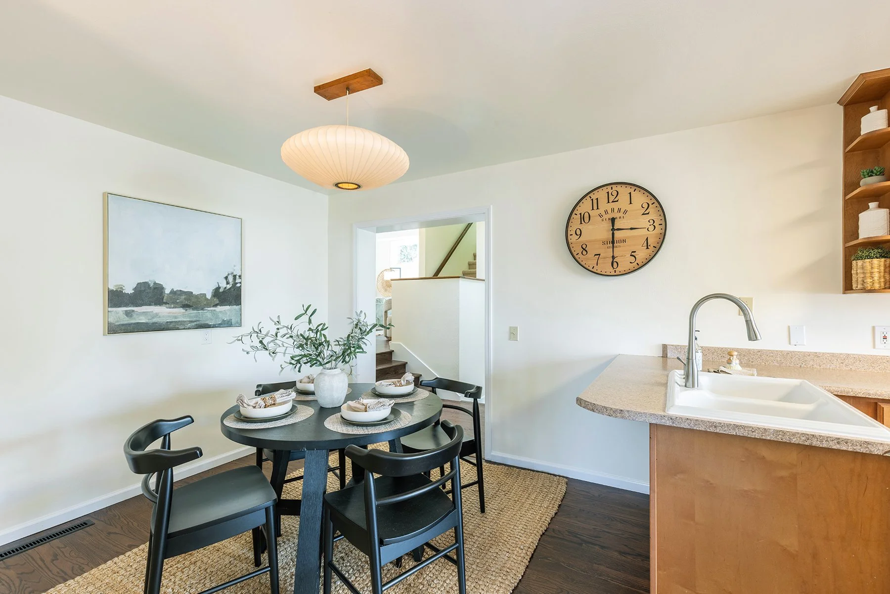 Dining area with a round table set with place settings, black chairs, a vase with greenery, and a hanging paper lantern. Adjacent to a kitchen counter with a sink, a large wall clock, a painting, and open shelves with decorative items.