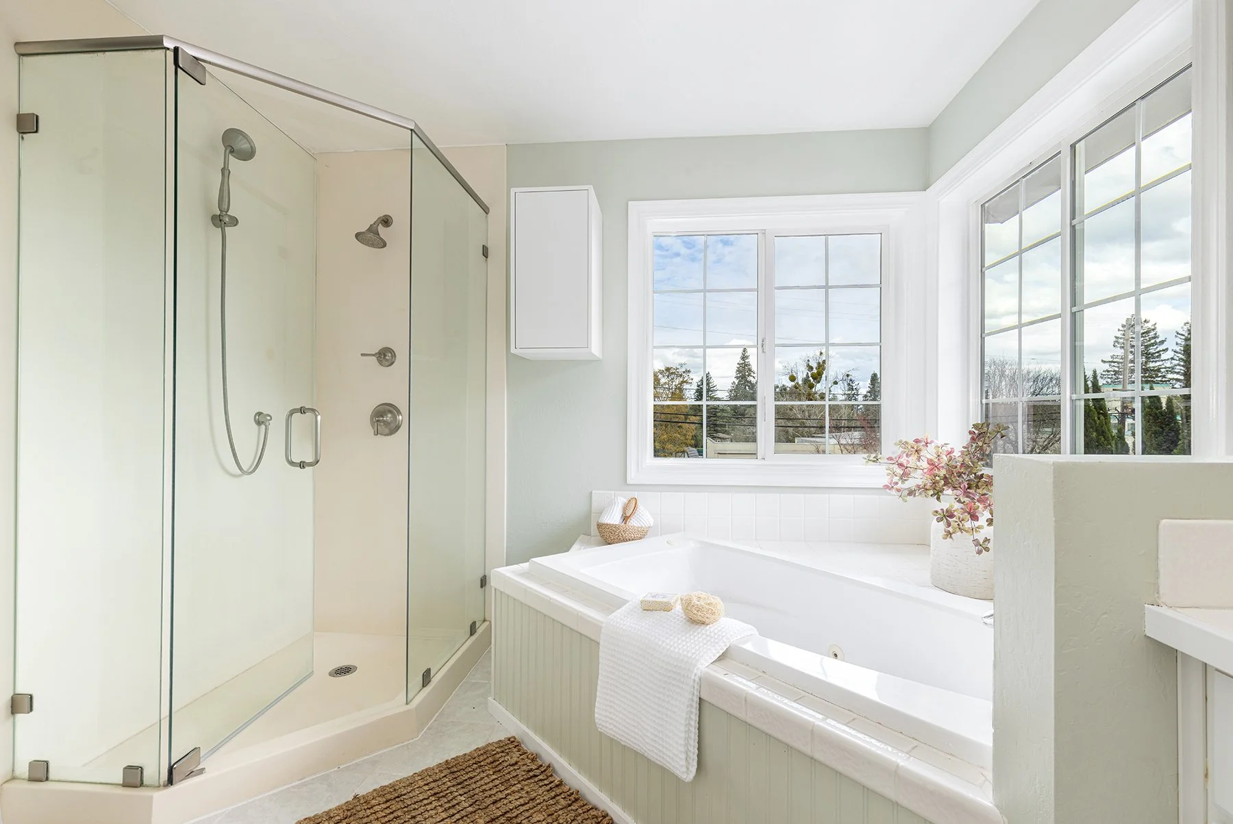Bright bathroom featuring a glass-enclosed shower, a white bathtub by large windows with a view of green trees, a potted plant, and a brown rug.