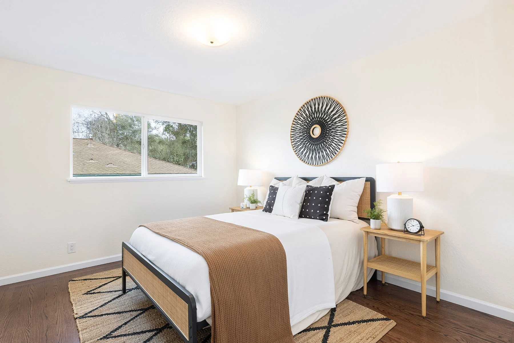 A bedroom with a large bed, white bedding, and black and white pillows. A wall decor above the bed, two bedside lamps, a clock, and a small plant on a wooden nightstand. Window showing a view of the rooftops and trees outside.