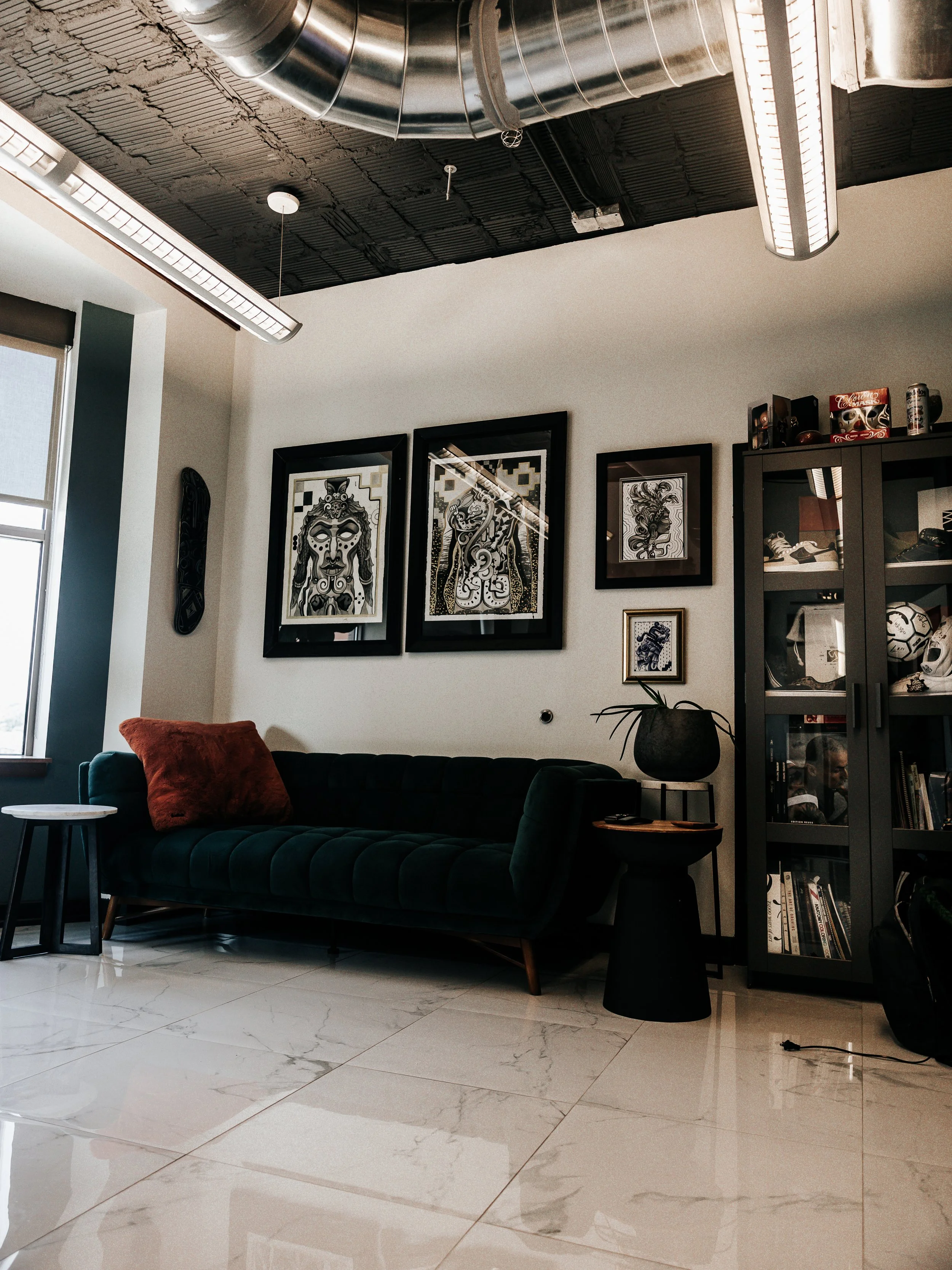 Modern living room with white tiled floor, black couch with a red throw pillow, black side table with a plant, a tall black cabinet with books and decorative items, black artwork frames on the wall, and industrial-style ceiling with exposed ductwork 