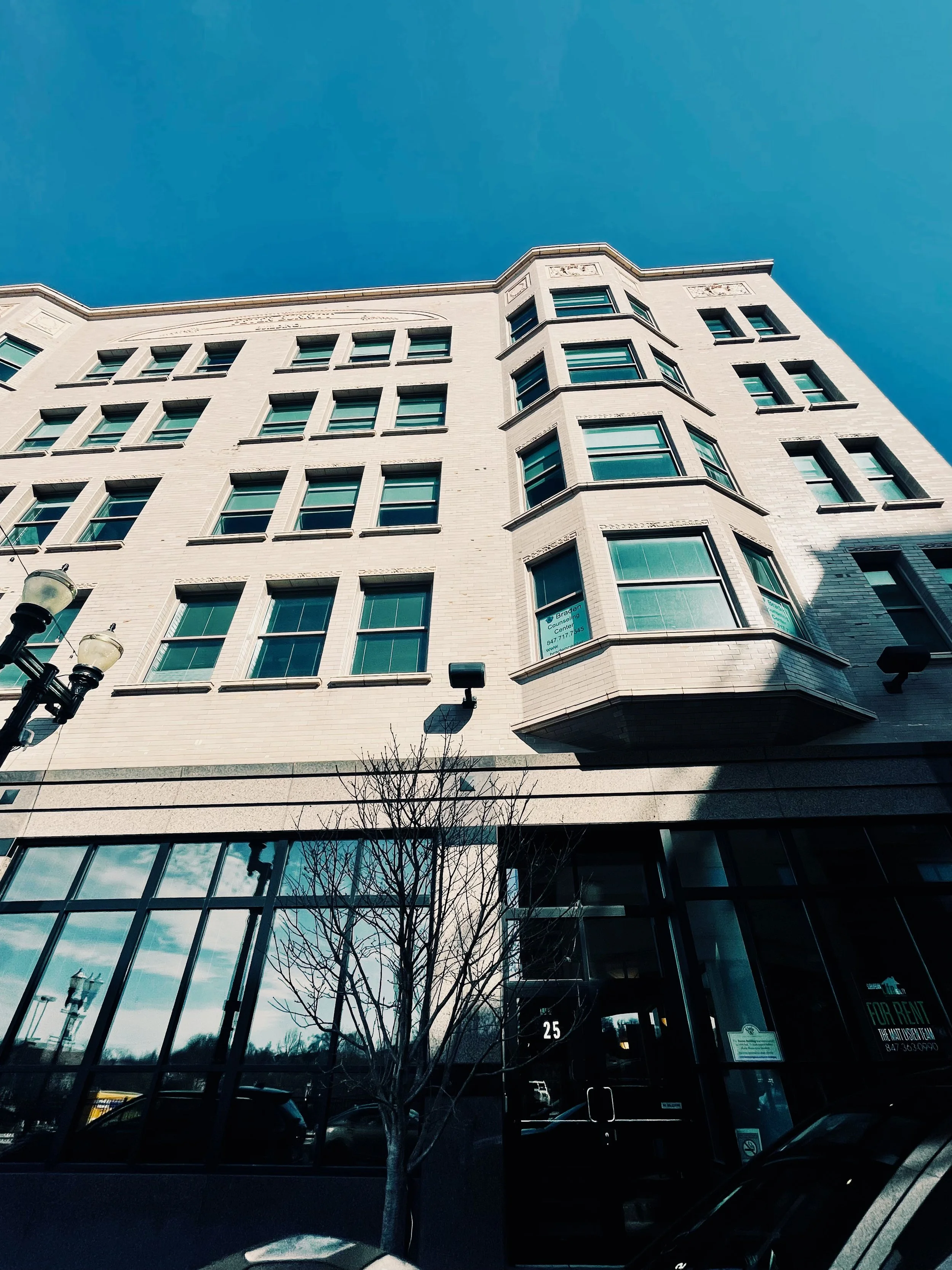 A tall, white brick building with multiple windows, some of which have signs indicating space for rent. The photo is taken from a low angle, showing a clear blue sky. There are leafless trees and cars parked in front of the building.